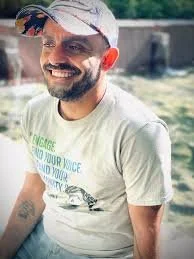 A smiling man with a beard wearing a white t-shirt and a baseball cap outdoors near a body of water.