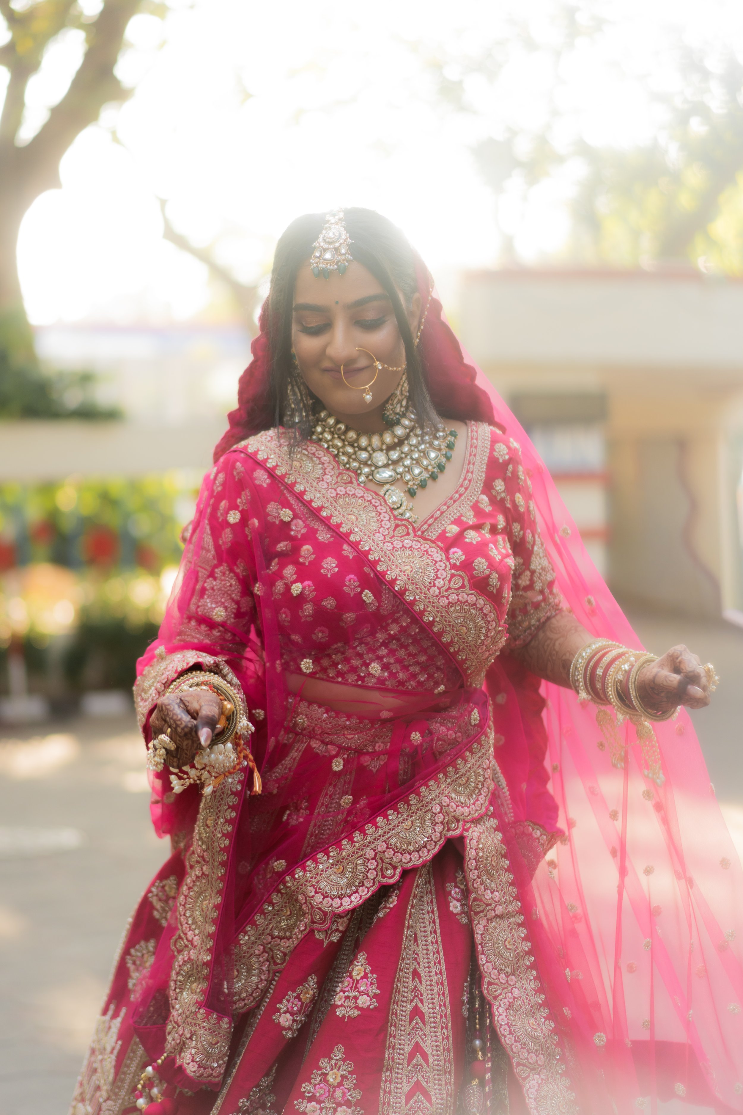 A woman dressed in a traditional Indian pink embroidered saree with gold and silver details, wearing statement jewelry including necklaces, earrings, a nose ring, bangles, and a headpiece, standing outdoors during daytime with sunlight behind her and trees in the background, appearing to be part of a wedding celebration or cultural event.