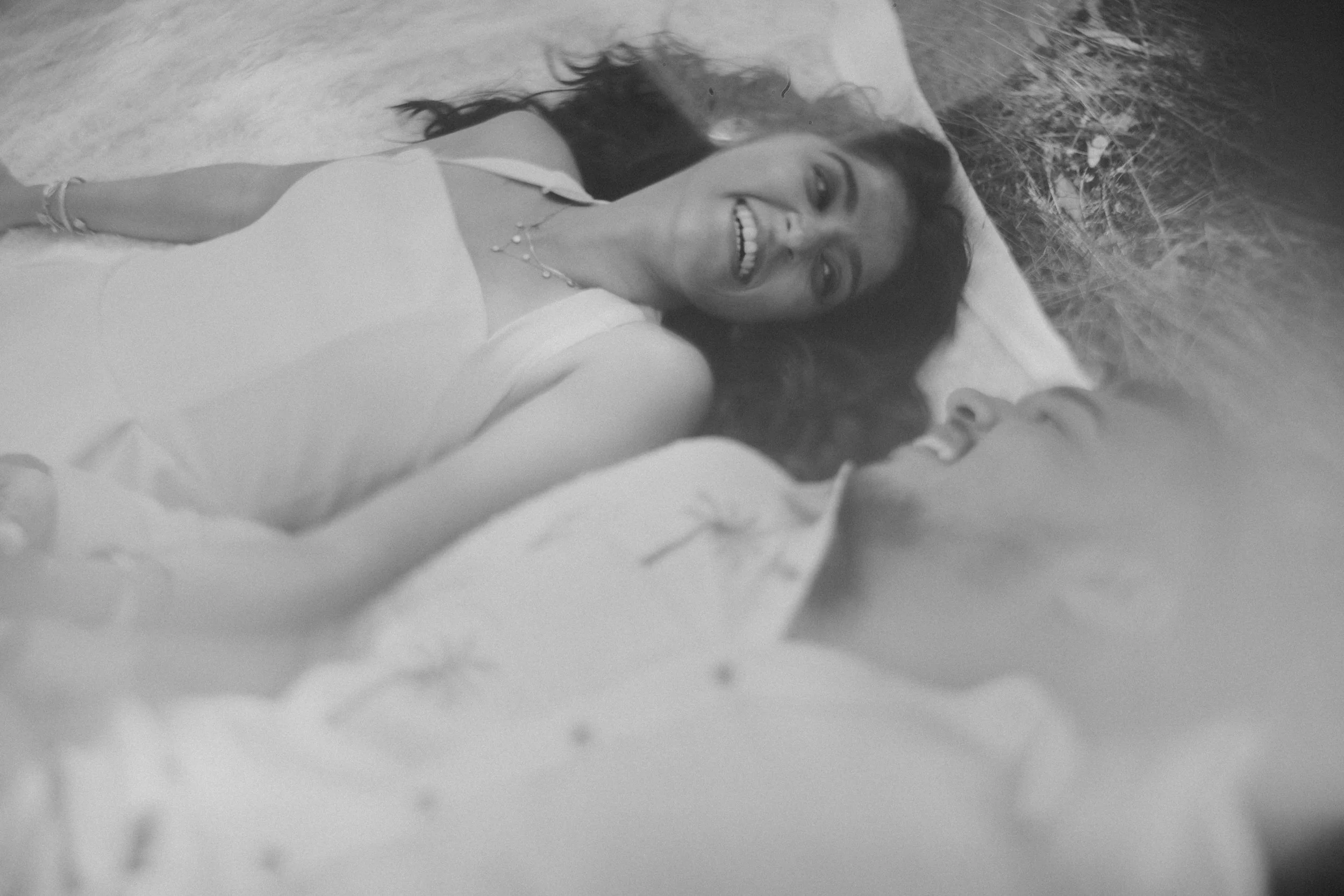 A young woman lying on her back in a natural outdoor setting, smiling and looking up at the camera. She has long hair and is wearing a sleeveless top, with jewelry visible on her wrist and necklace.