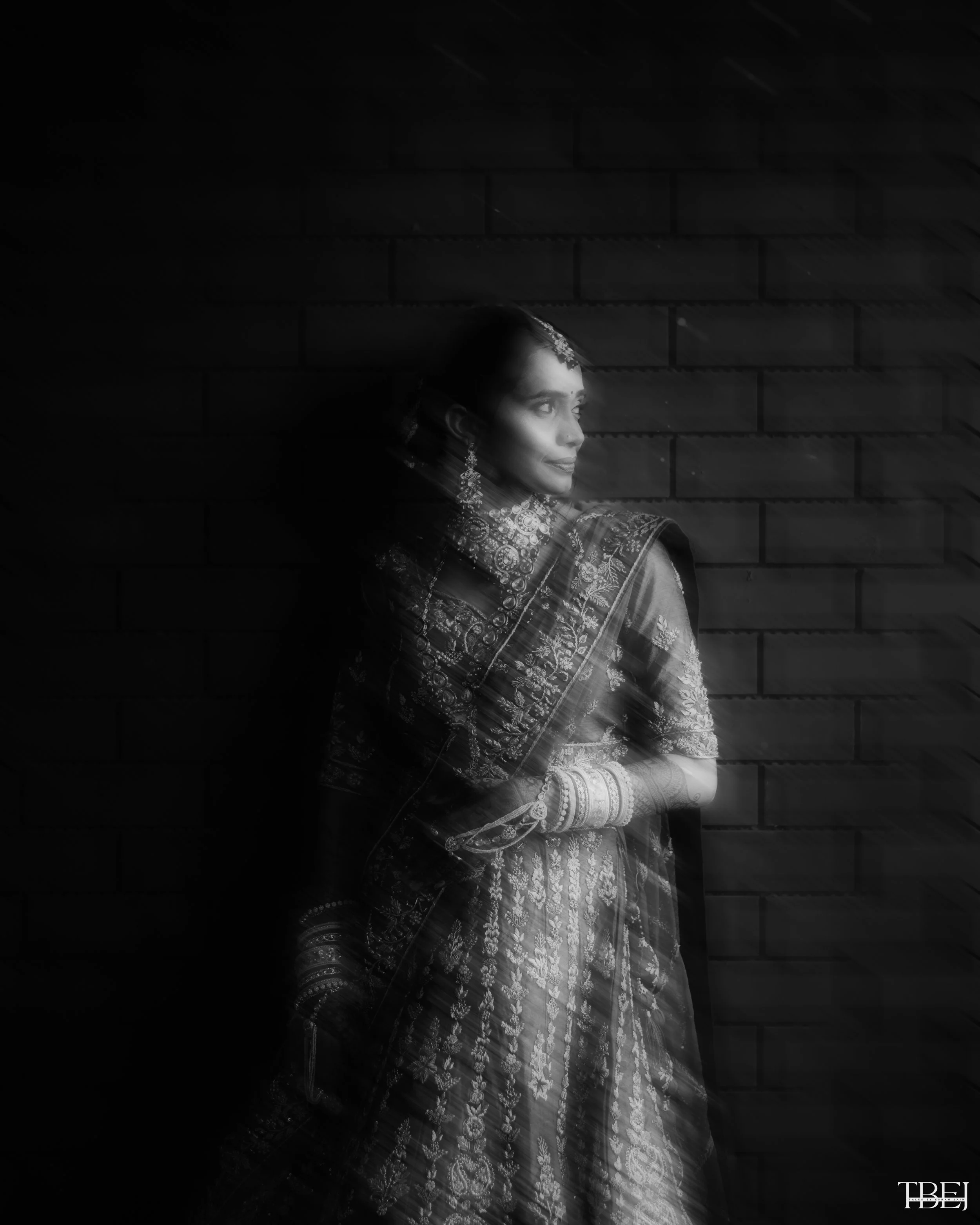 A woman in traditional Indian attire with jewelry, gazing to the side with a soft expression, standing against a brick wall with light streaks passing through.