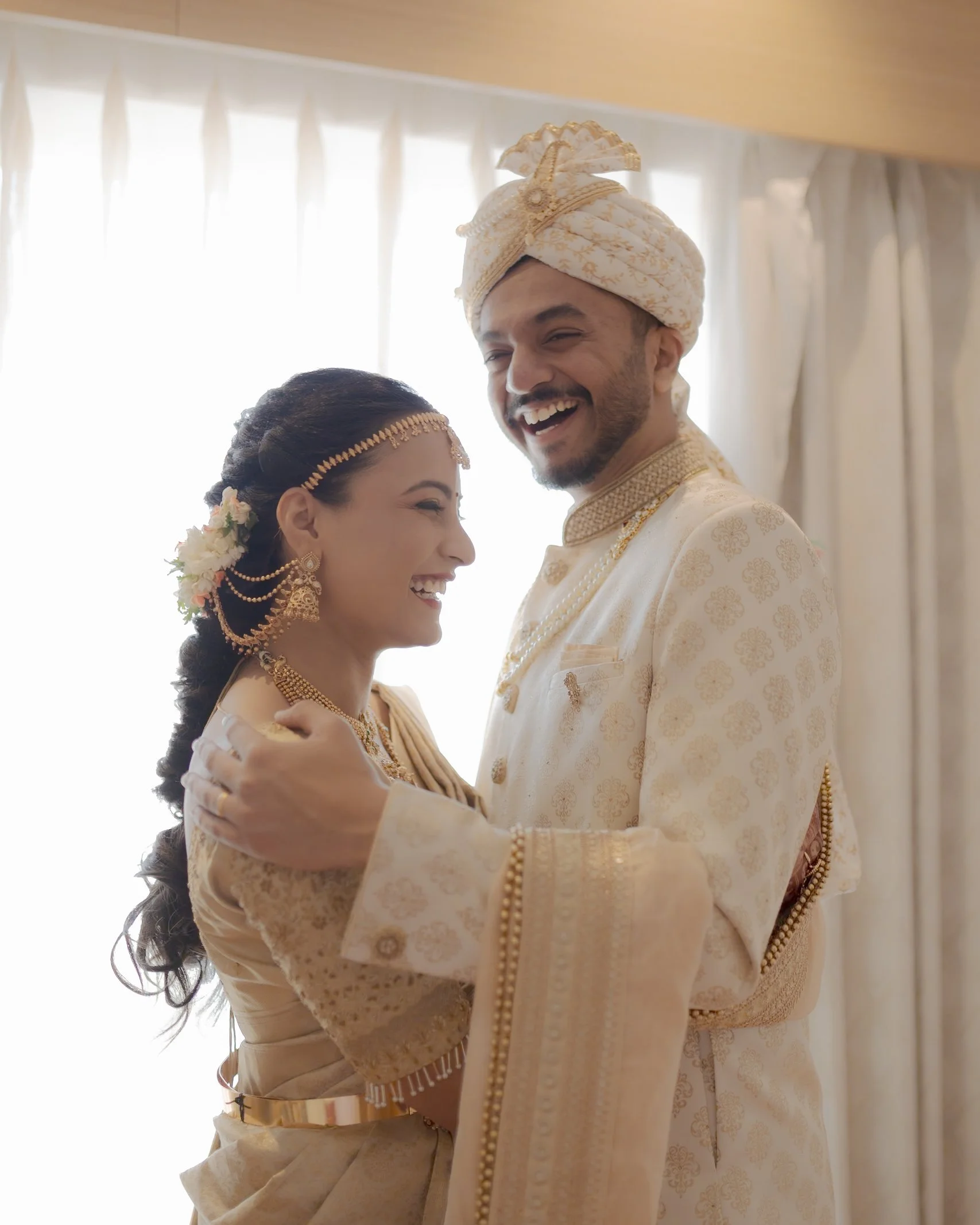 A bride and groom in traditional Indian wedding attire, smiling and holding each other indoors with curtains in the background.
