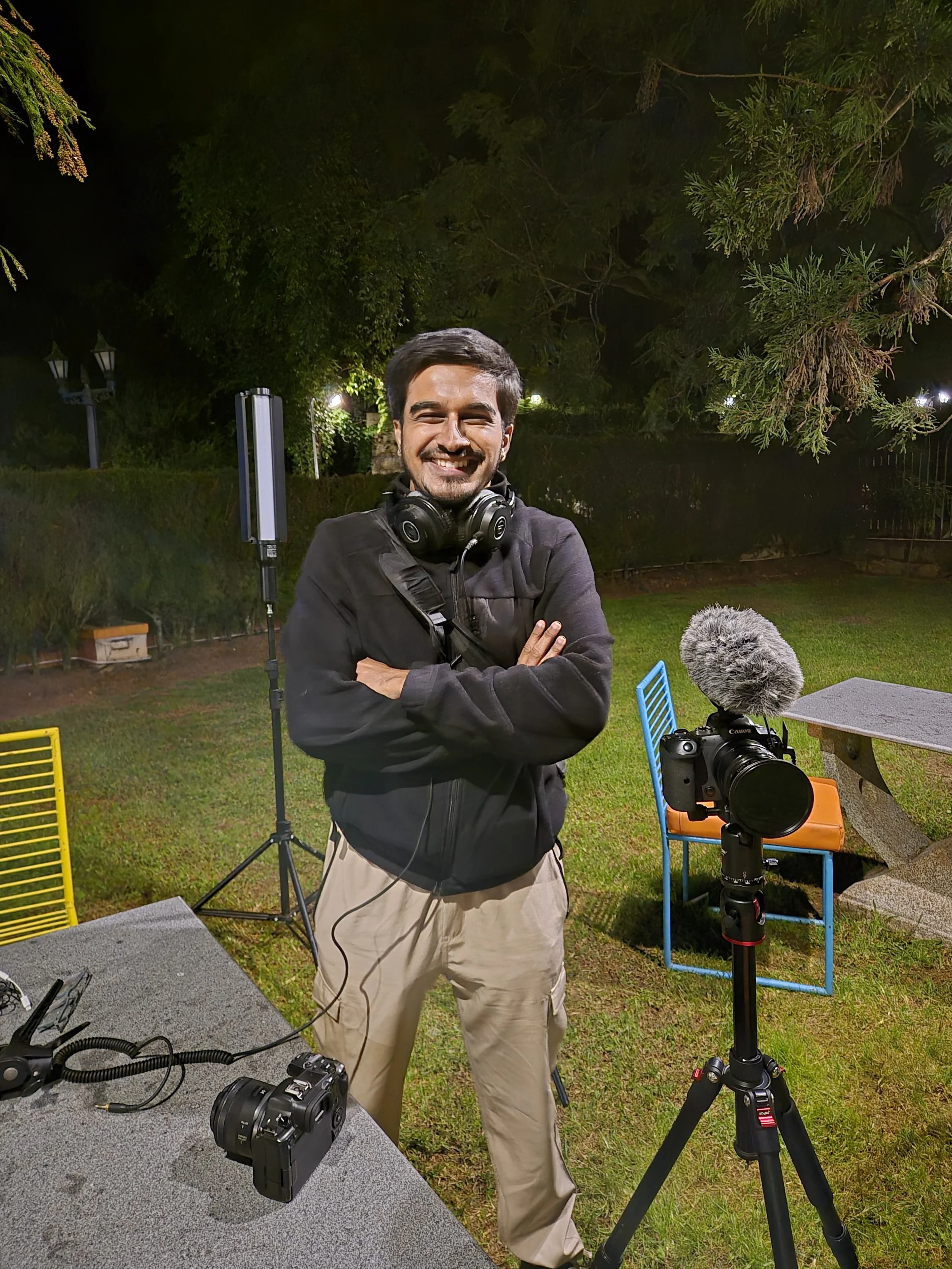 A man smiling and standing outdoors at night, wearing a black jacket with a camera around his neck, surrounded by photography equipment including a camera on a tripod, a large microphone, and additional cameras on a table.