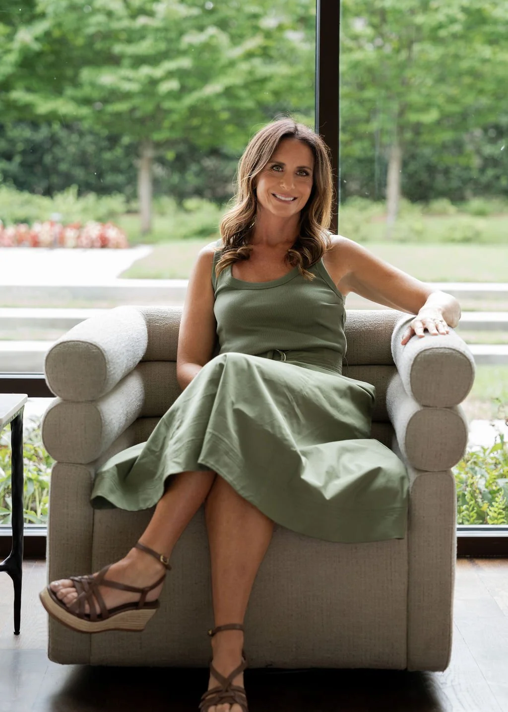 Brittany sits on a beige armchair in a room with large windows overlooking a green outdoor landscape, smiling and wearing an olive green sleeveless top, a matching skirt, and brown wedge sandals.