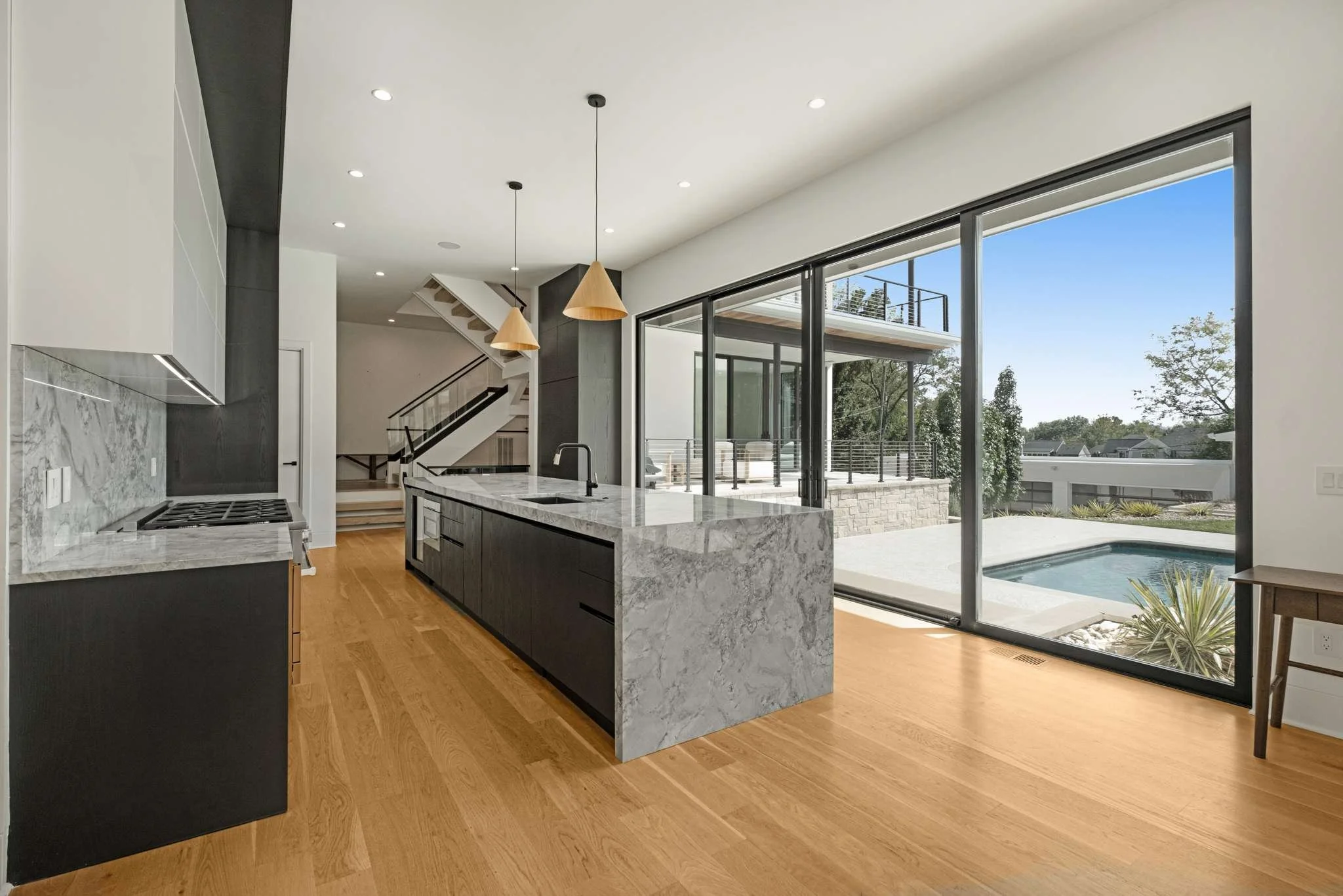 Modern kitchen with marble island and large sliding glass doors leading to outdoor patio and pool area, with staircase and outdoor seating visible.
