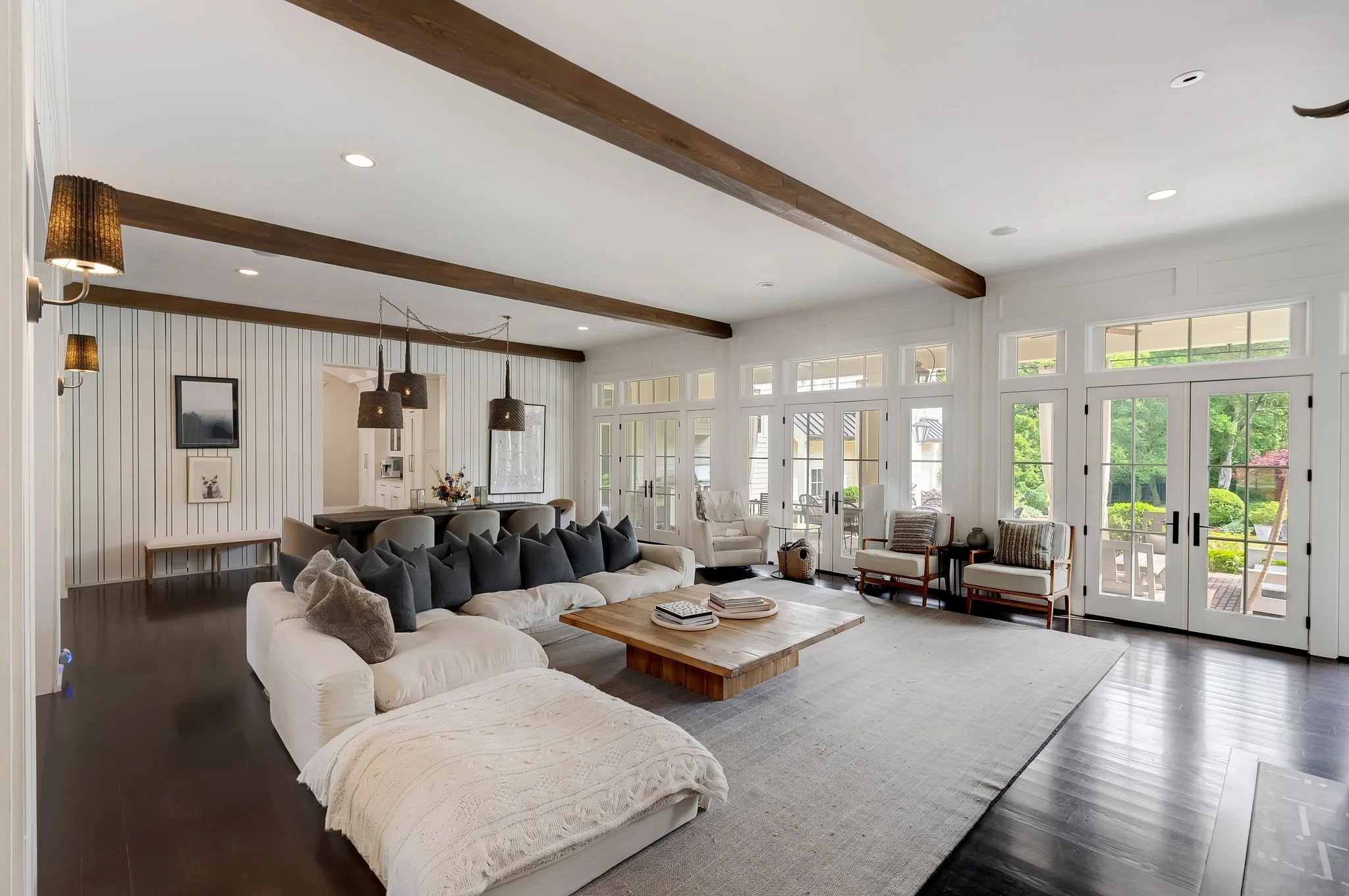 A spacious living room with large windows, white walls, and dark wood flooring. The room features a white sectional sofa with dark pillows, a wooden coffee table, armchairs, and various decorations including framed pictures and lamps.