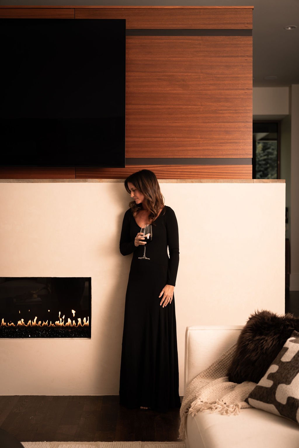 Brittany wears a black dress holding a glass of red wine, standing next to a modern fireplace with a minimalist design, in a stylish, contemporary living room.