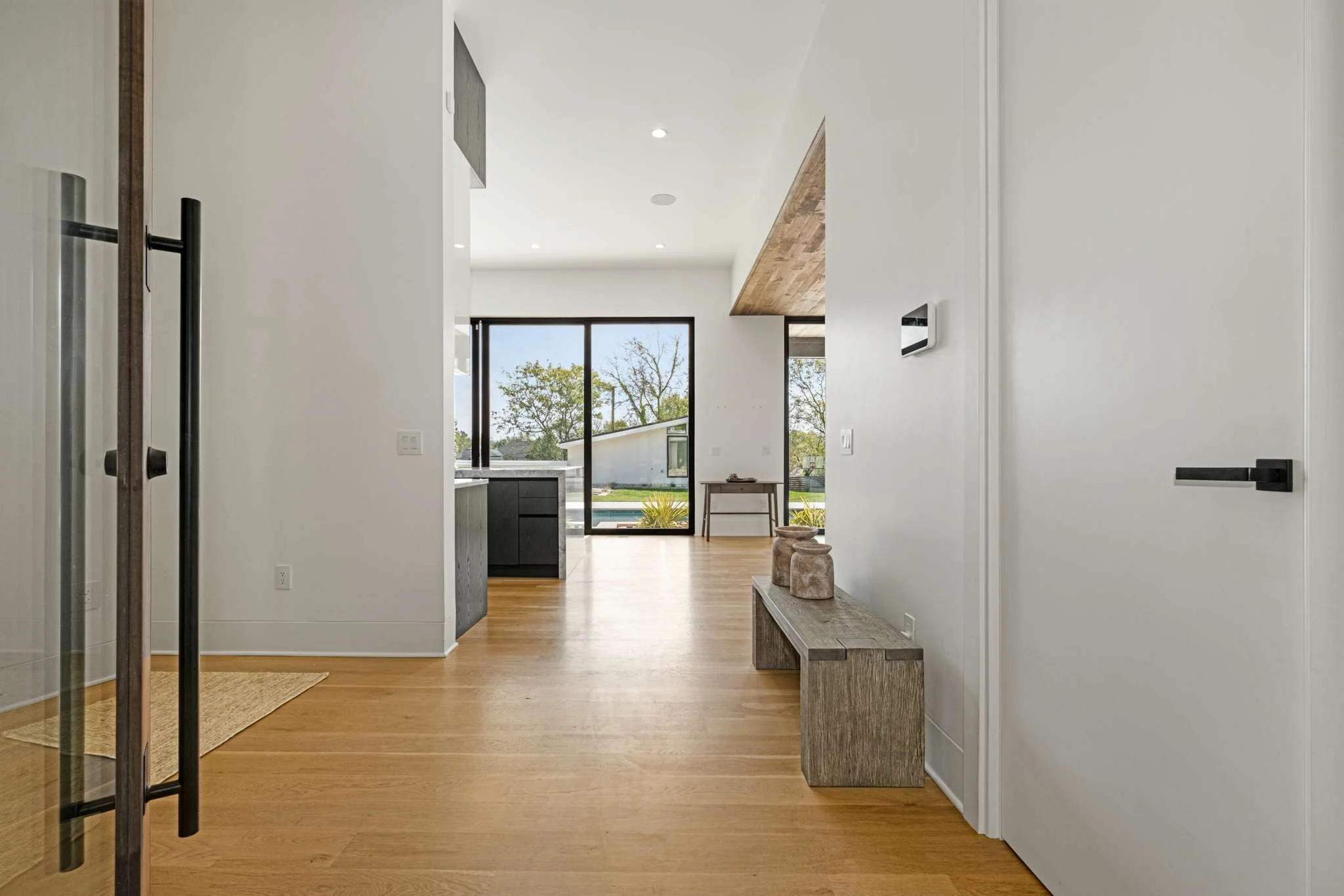 Modern interior entryway with light wooden flooring, white walls, a black door handle, and a view into a bright living space with large windows and trees outside.