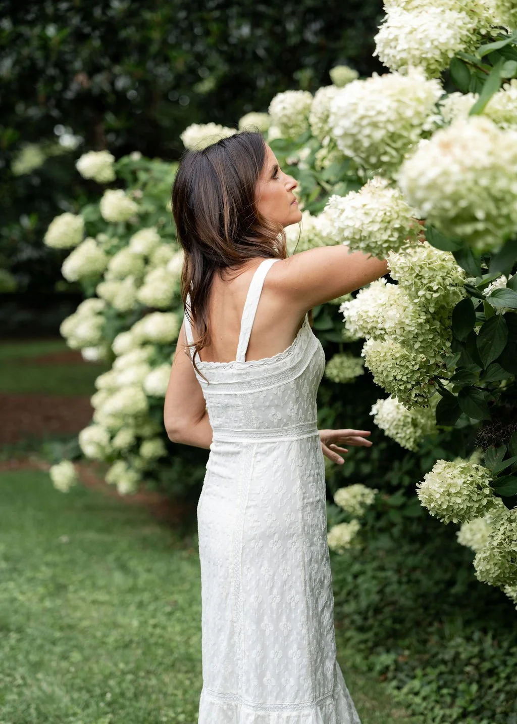 Brittany stands in a garden with large white hydrangea flowers, gently touching the blooms while facing sideways.