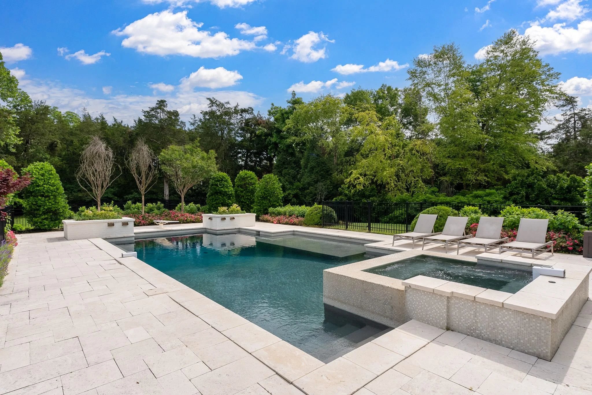 A backyard with a swimming pool surrounded by white stone tiles, poolside lounge chairs, lush green trees, and a bright blue sky with fluffy clouds.