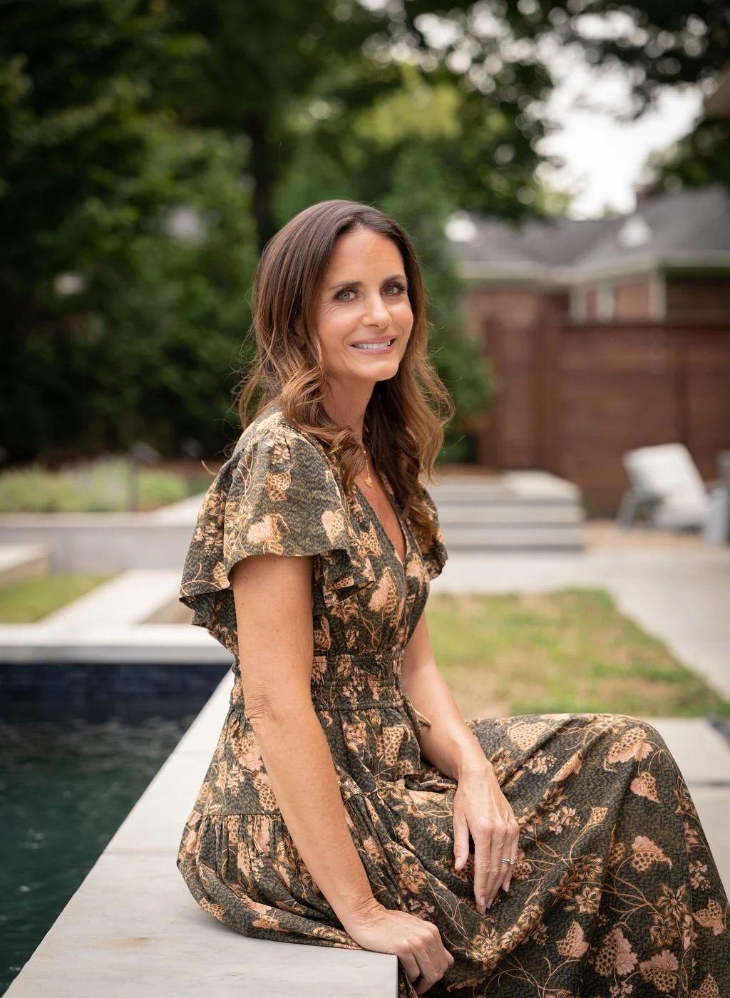 Brittany Sircy wears a floral dress, sitting on the edge of a pool in an outdoor setting with greenery and a brick house in the background.