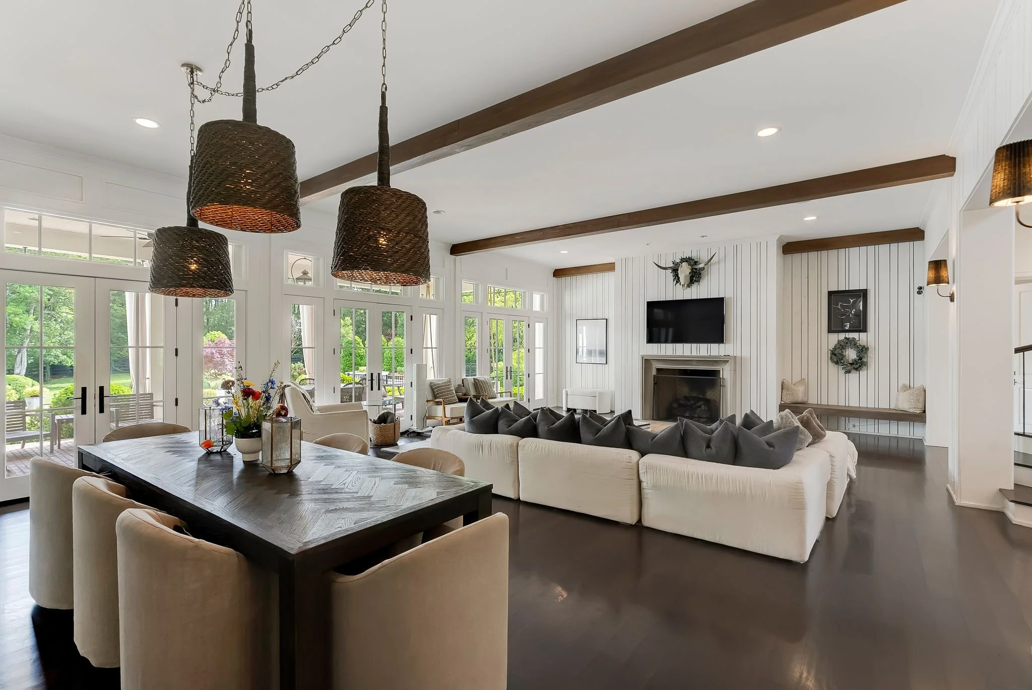 A spacious living room with white walls, wooden ceiling beams, and large windows with glass doors leading to an outdoor area. The room features a white sectional sofa with dark gray pillows, a wooden dining table surrounded by beige chairs, and black woven pendant lights hanging from the ceiling. There is a fireplace with a decorative animal skull and wreath above it, and framed artwork on the wall.