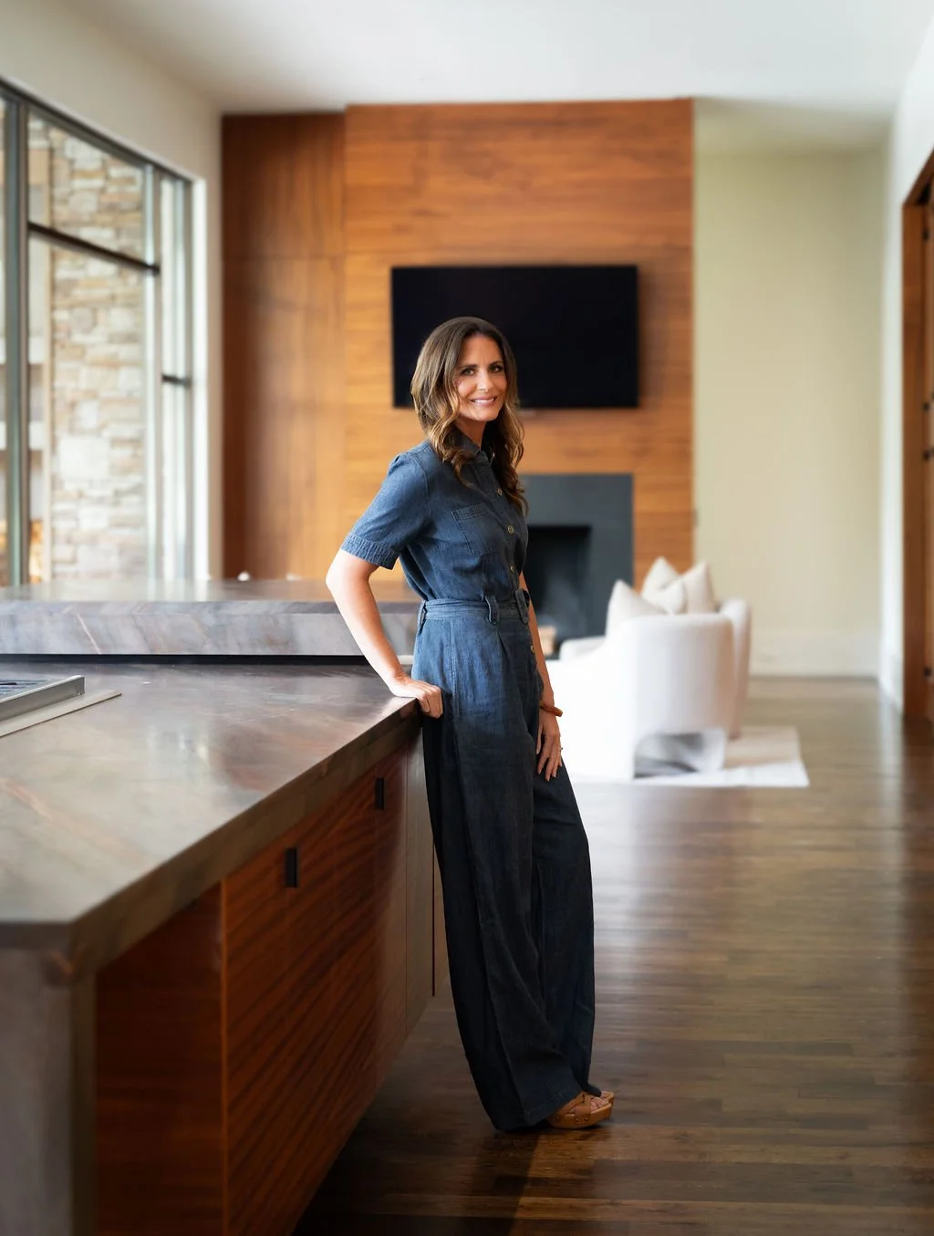 Brittany, of Sircy & Co., wears a dark blue jumpsuit standing in a modern living room with wood-paneled wall and flat screen TV