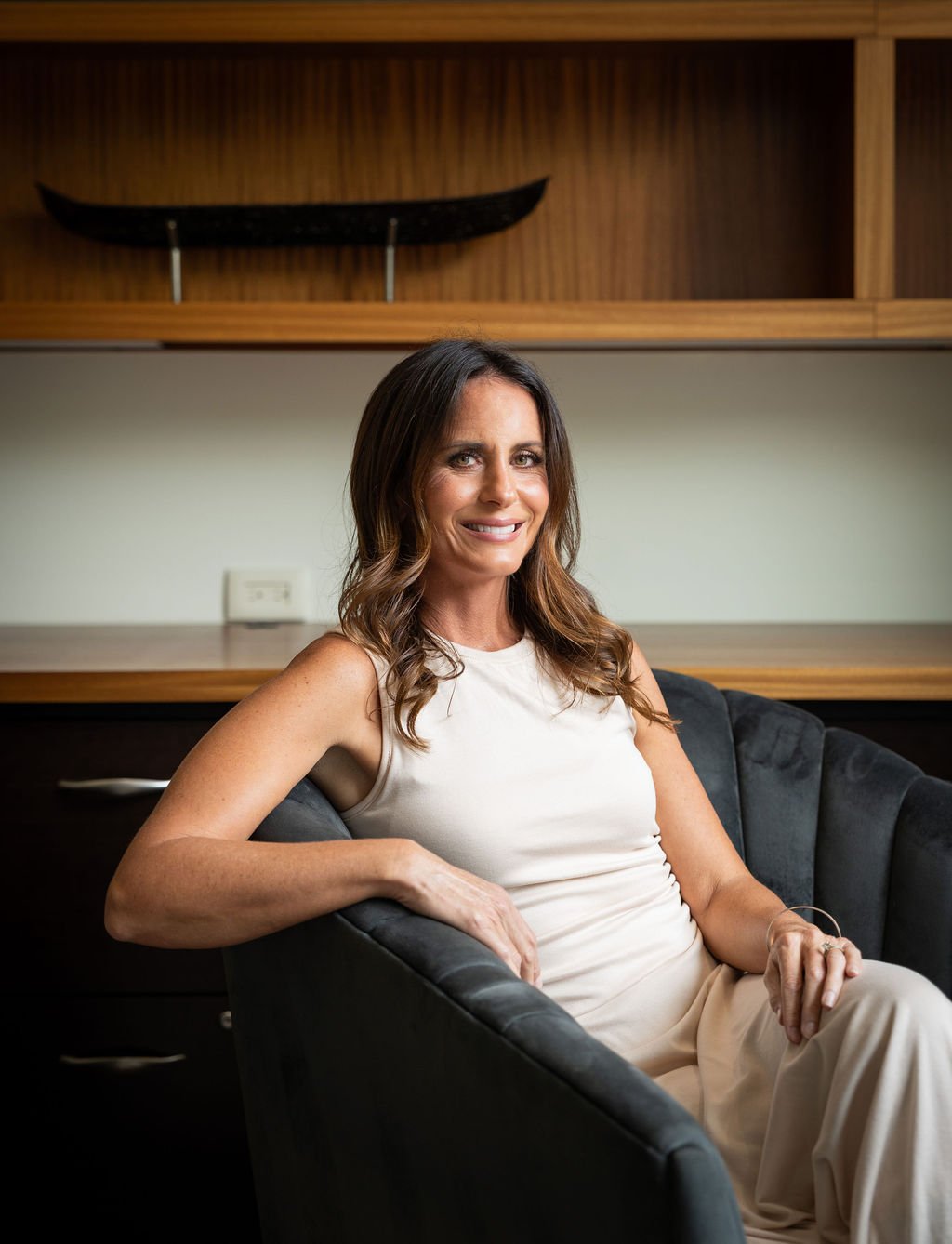 Brittany sits in a black armchair in a room with wood-paneled shelves and a decorative black skateboard on the wall behind her. She is smiling, wearing a sleeveless white top and beige pants.