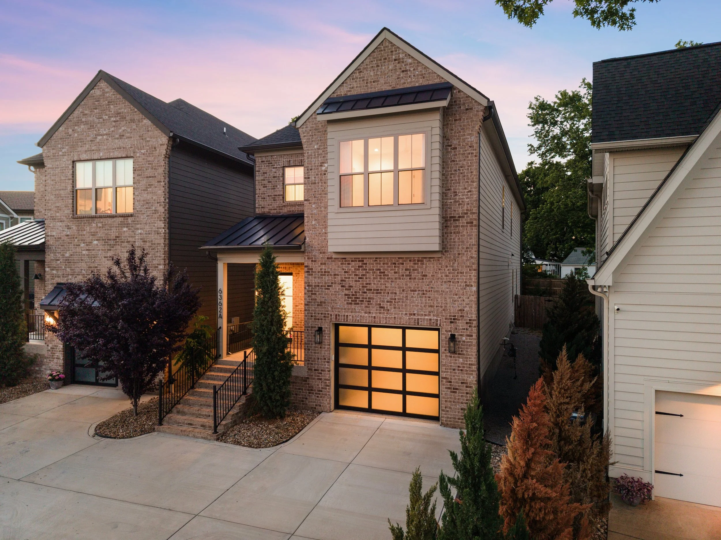 A modern multi-story house with brick and siding exterior, large illuminated windows, and a black garage door, at dusk with a pink and blue sky backdrop.