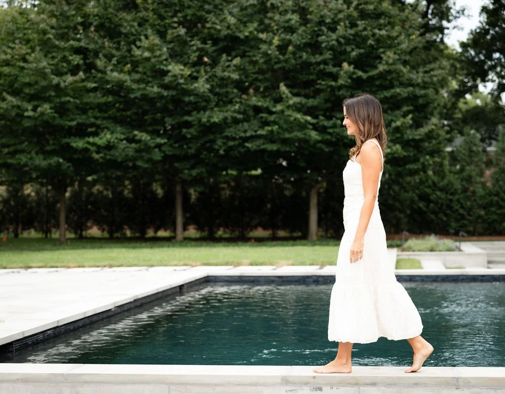 Brittany Sircy wears a white dress and walks barefoot on the edge of a pool outdoors.