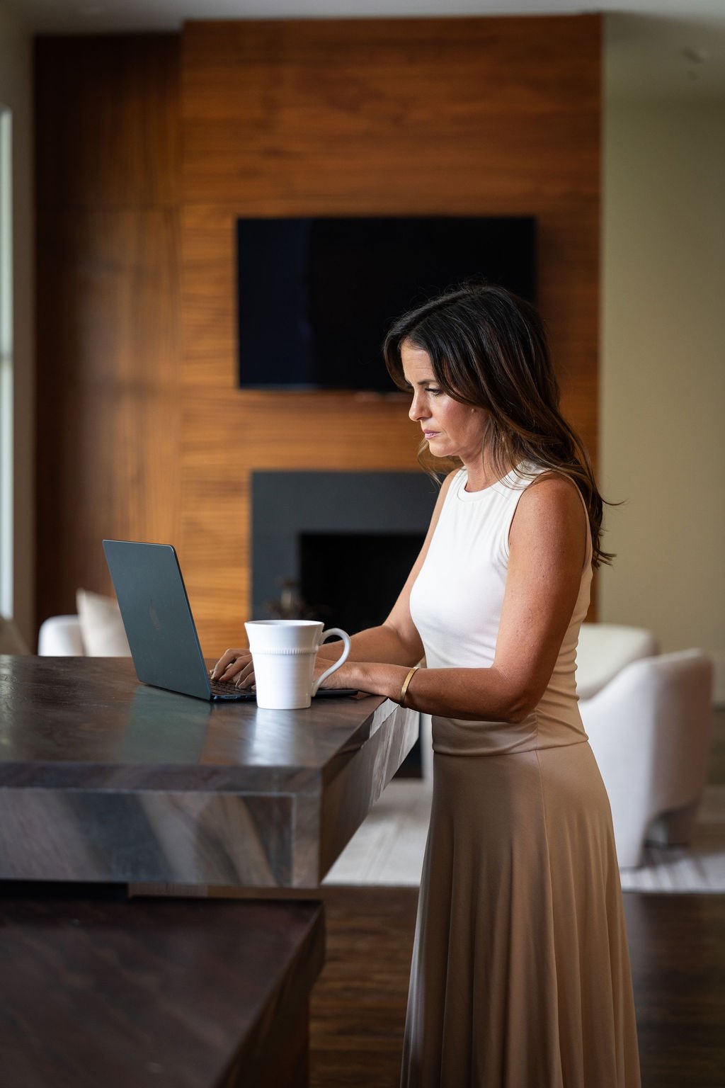 Brittany, in a sleeveless white top and beige skirt works on a laptop at a wooden kitchen island, with a white mug beside her, in a modern home with wood-paneled walls and a large flat-screen TV in the background.