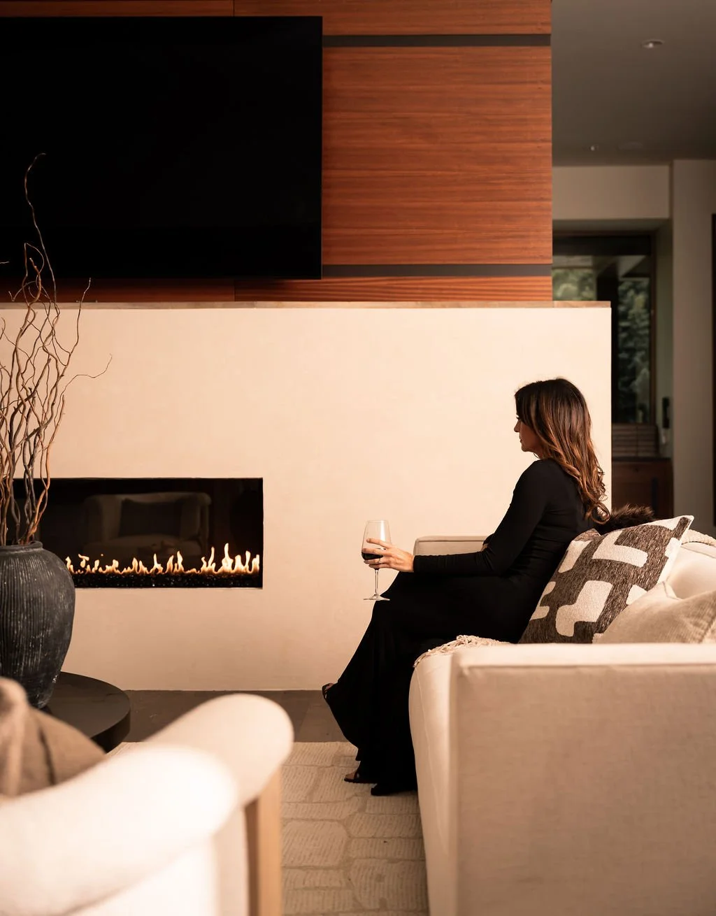 Brittany, wearing a black dress sits on a beige sofa holding a glass of red wine, near a modern fireplace in a stylish living room.