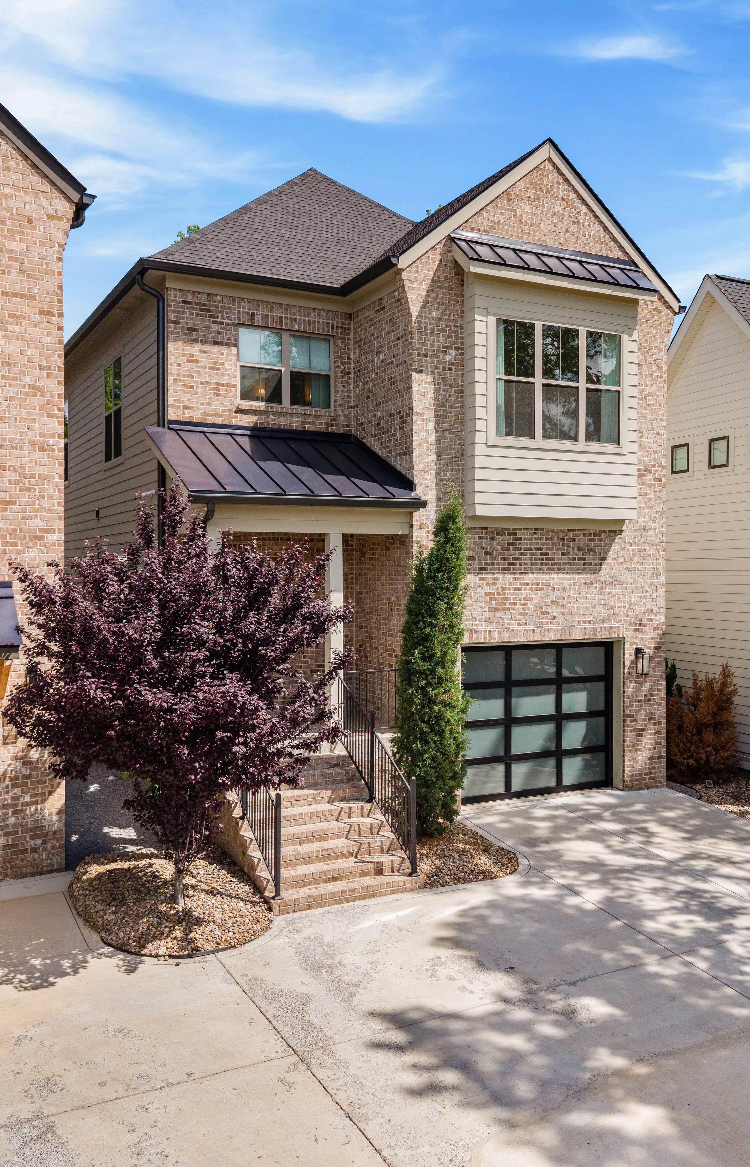 A modern two-story house with brick and siding exterior, large front window, black metal roof, garage with black panel doors, stone stairs, a purple-leafed tree, and minimal landscaping. Clear blue sky overhead.