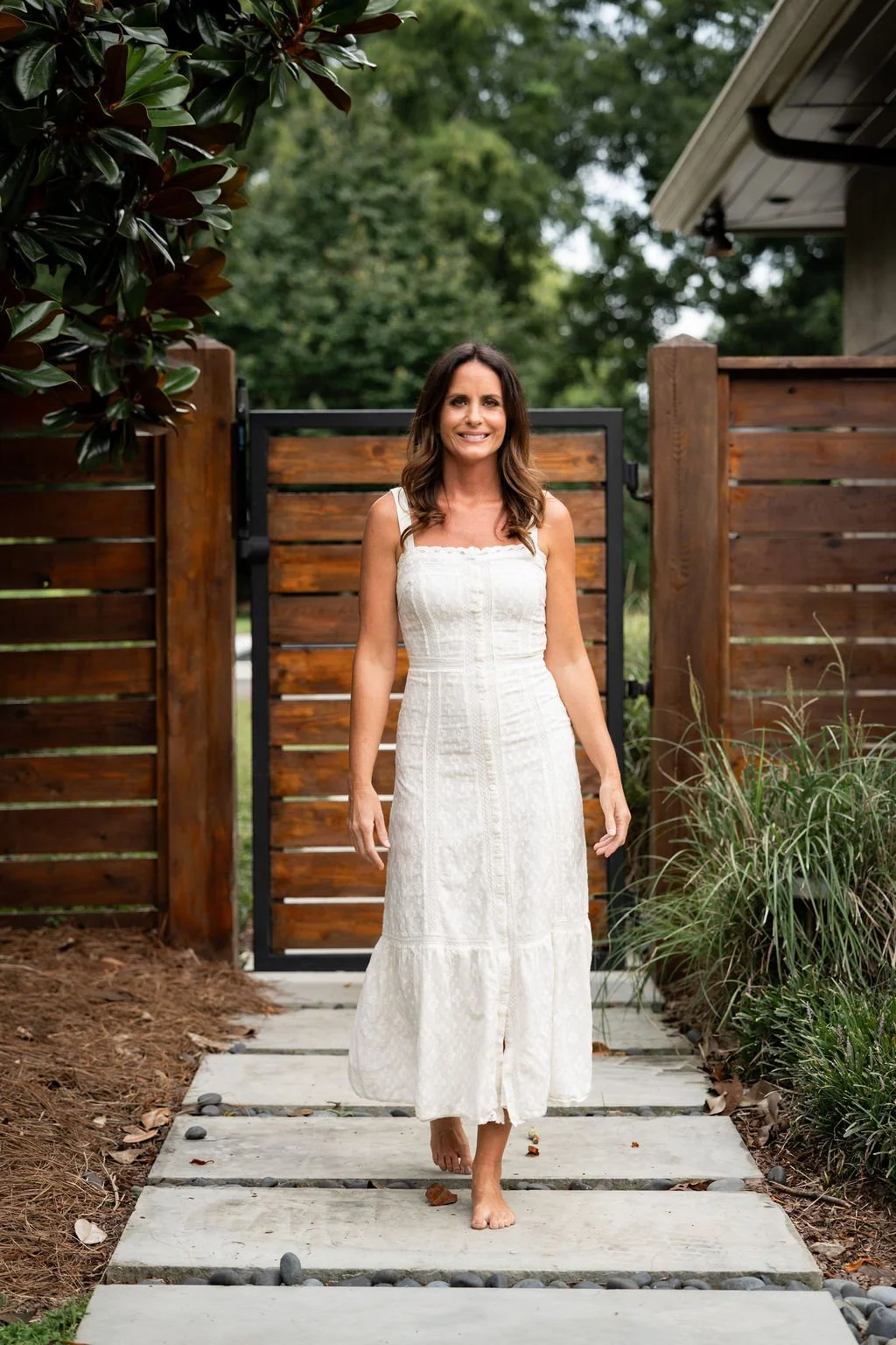 Brittany, in a white dress, walks barefoot on a stone pathway outdoors, with a wooden fence and greenery in the background.