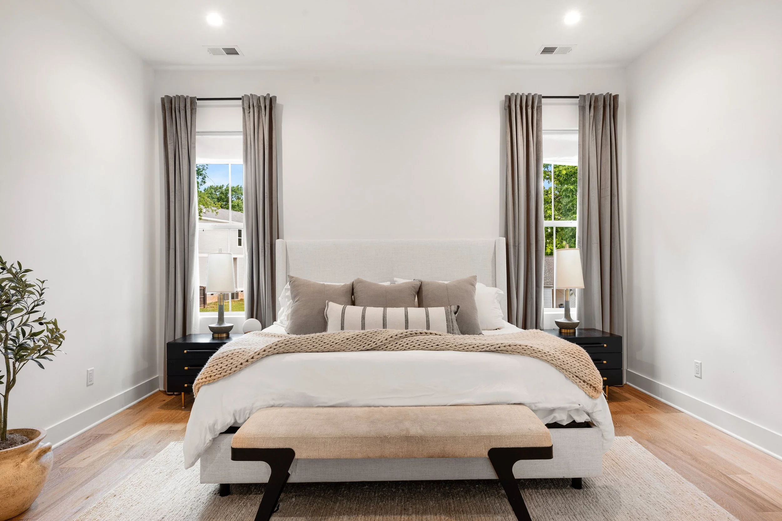 Modern bedroom with white walls, two tall windows with gray curtains, a bed with white linens, gray and striped pillows, a beige knitted blanket, black side tables with white lamps, a beige bench at the foot of the bed, a potted plant on the left, and hardwood floors.