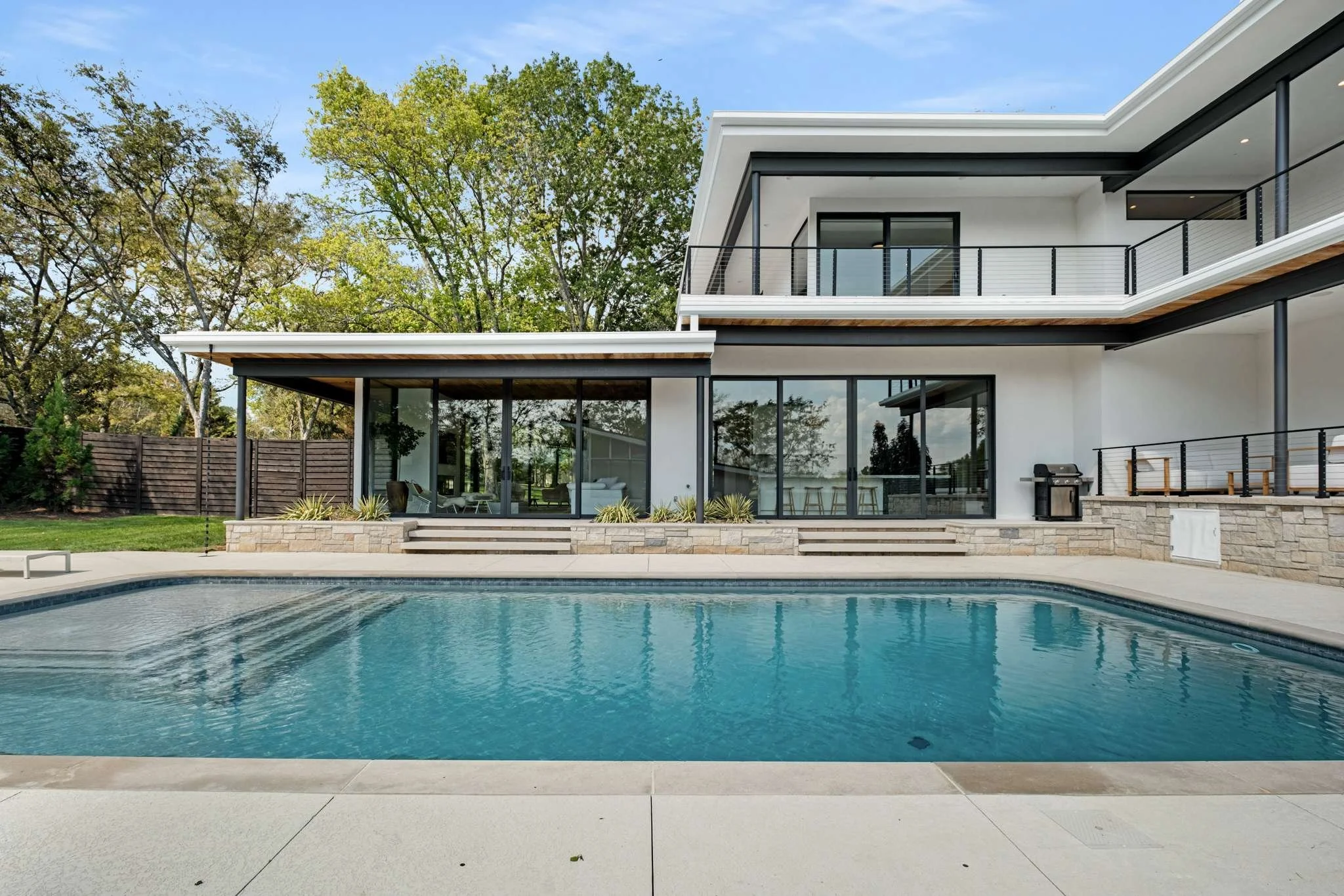 Modern house with large glass windows and balcony, backyard with swimming pool surrounded by patio and greenery, blue sky in background.