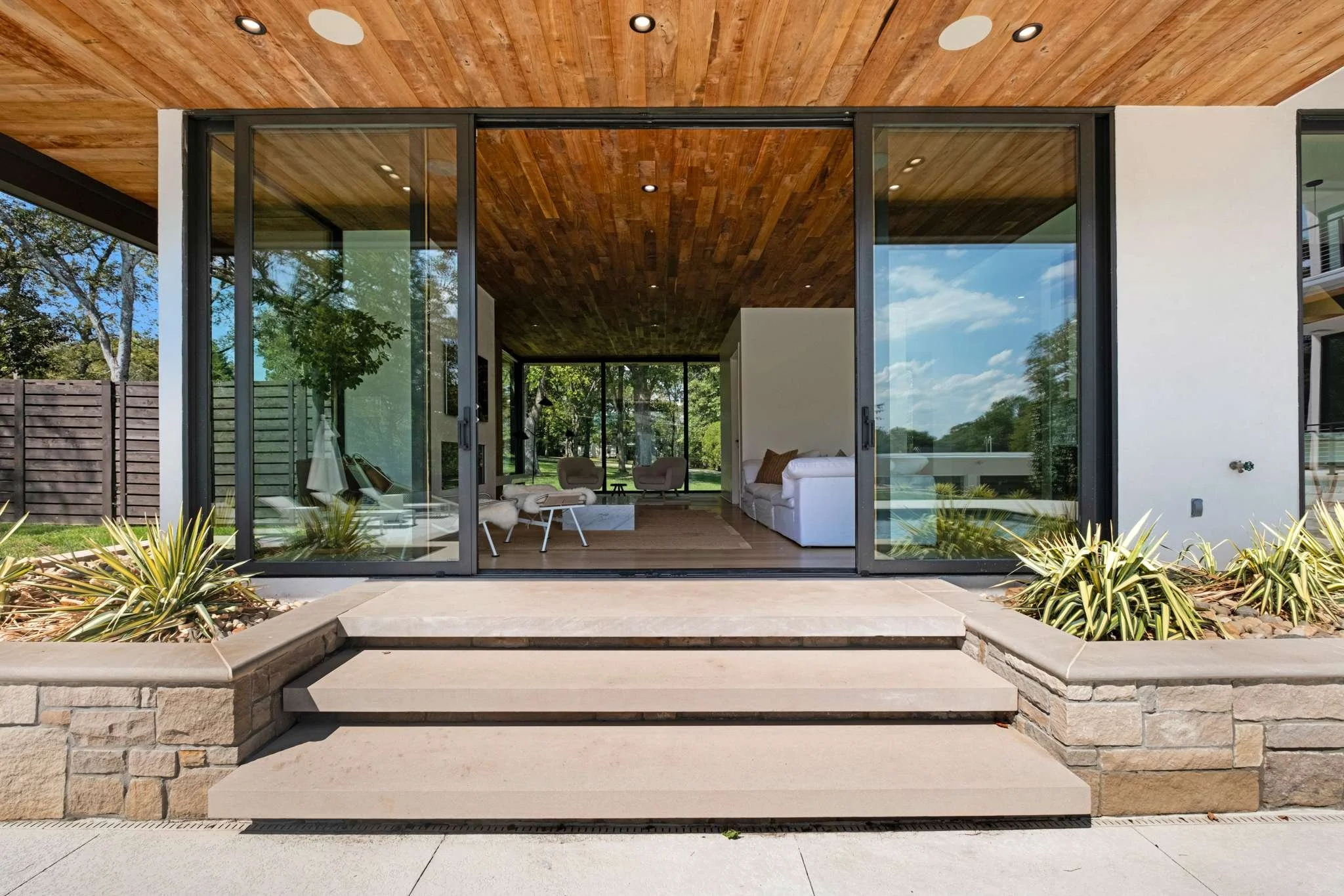 Front of a modern house with large glass sliding doors, a wooden ceiling, and stone steps leading inside