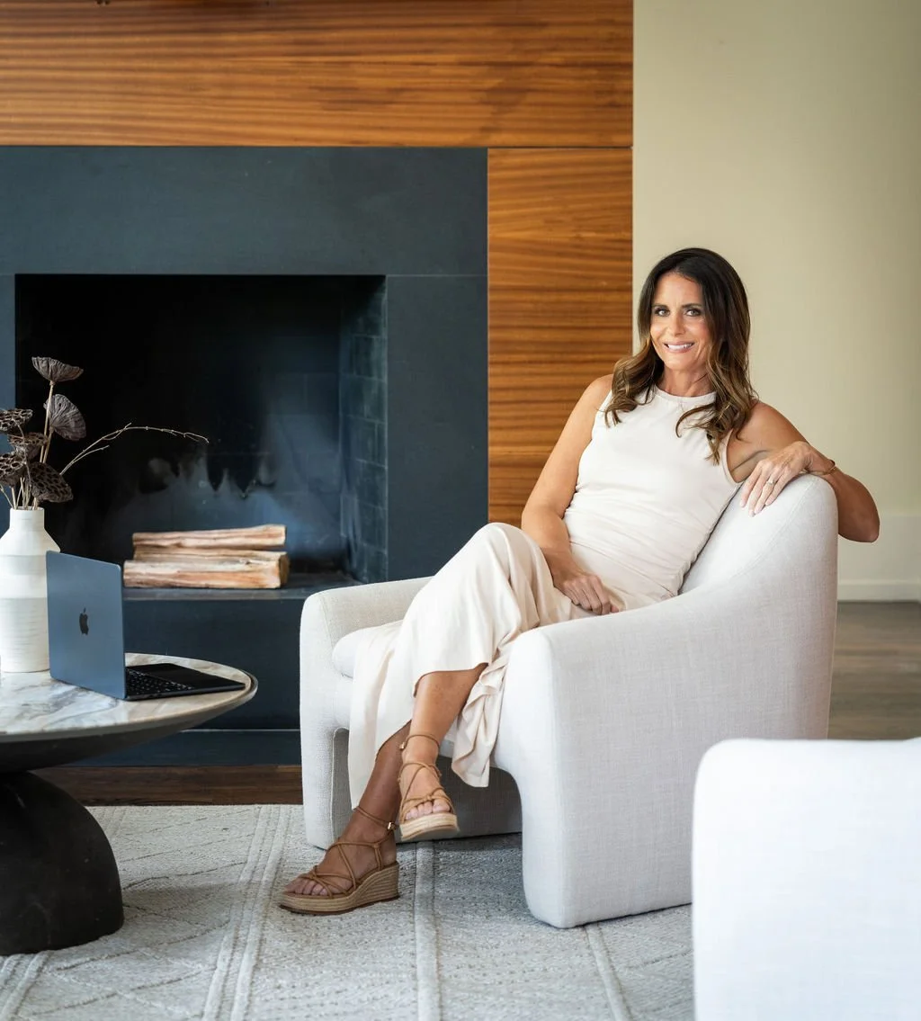 Brittany sits in a white armchair in a modern living room with wood and black fireplace surround, a marble top table with a laptop, and decorative plants.