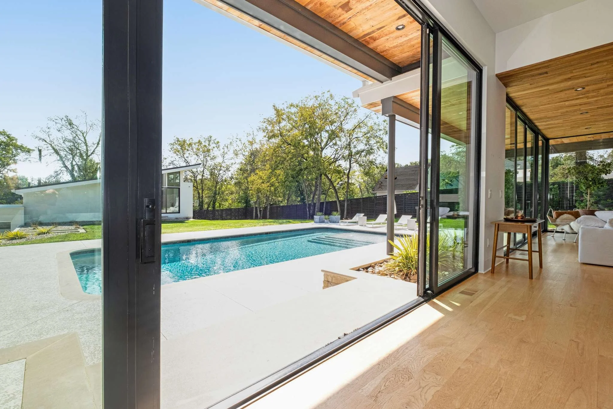 Living room with large sliding glass doors opening to a backyard with a pool, surrounded by trees and patio furniture, with a wooden ceiling and modern interior decor.