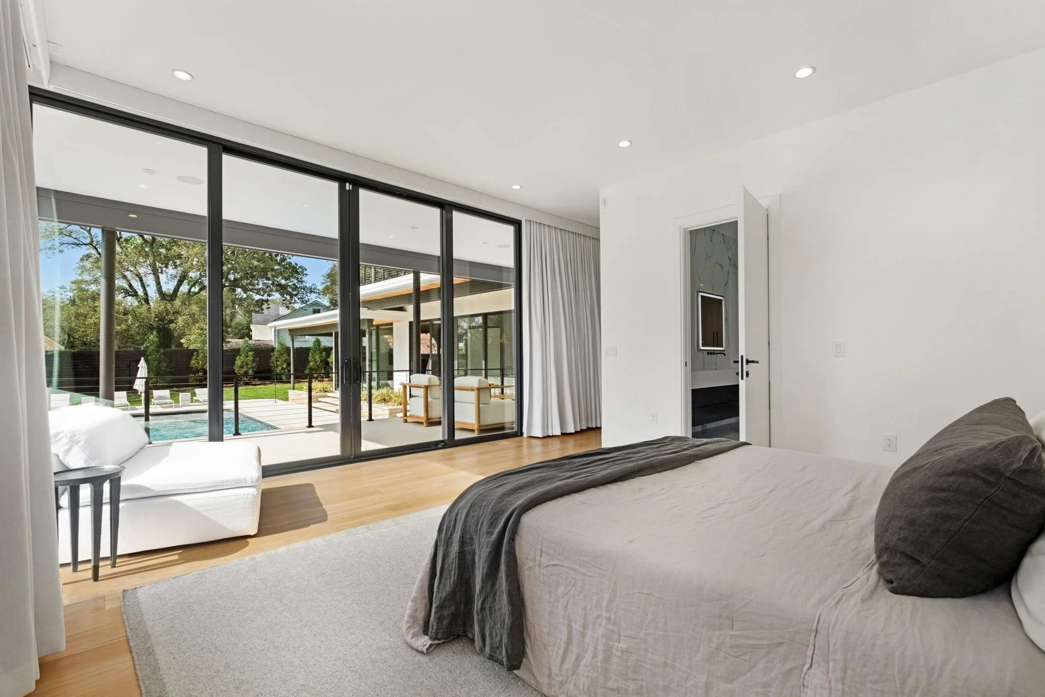 A modern bedroom with a large sliding glass door opening to a backyard with a pool, trees, and outdoor seating. The room has white walls, a grey bedspread, a white armchair, a small side table, and cream-colored curtains.