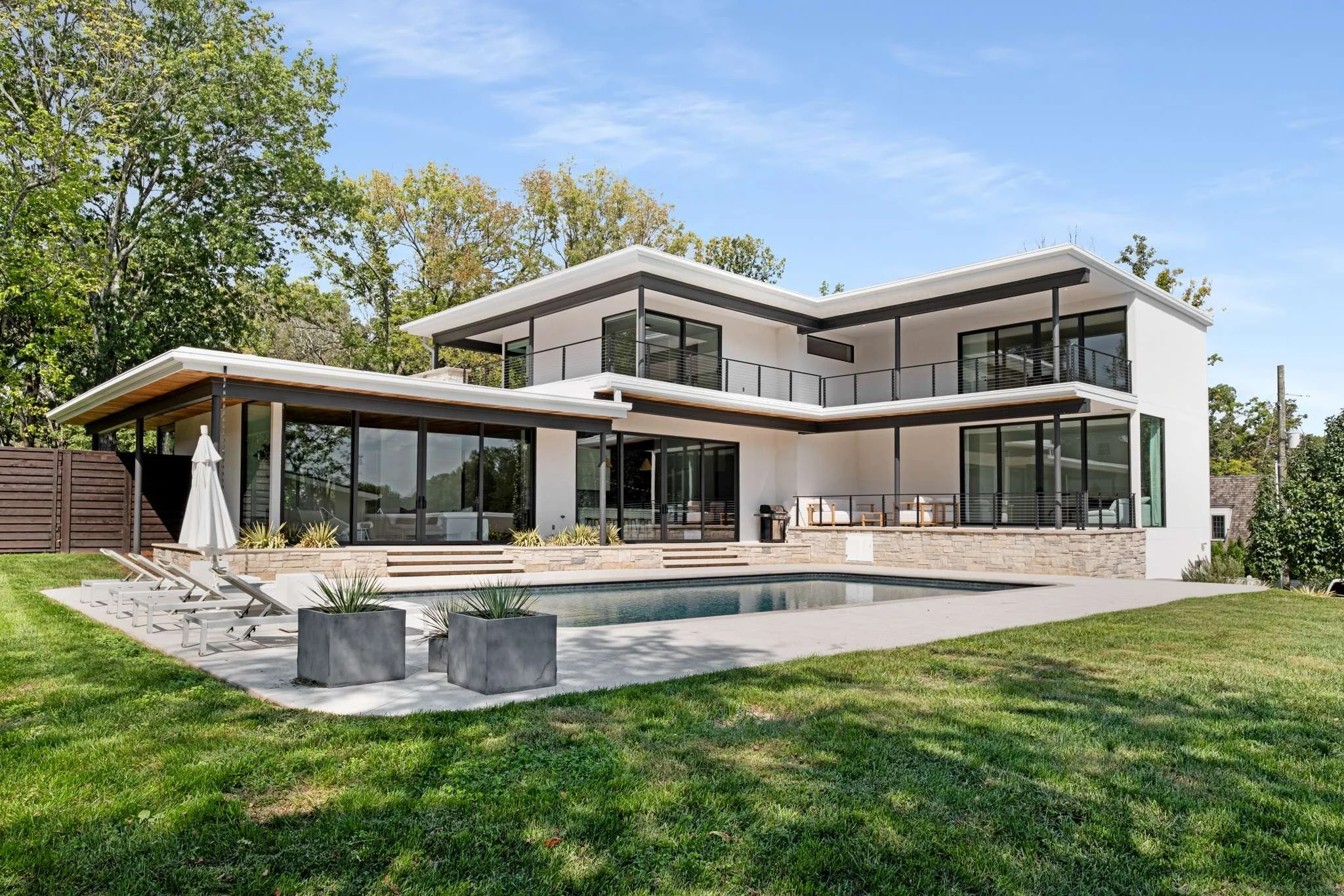 Modern two-story house in Tennessee with large glass windows, a balcony, and an outdoor swimming pool, surrounded by green grass and trees under a clear blue sky.