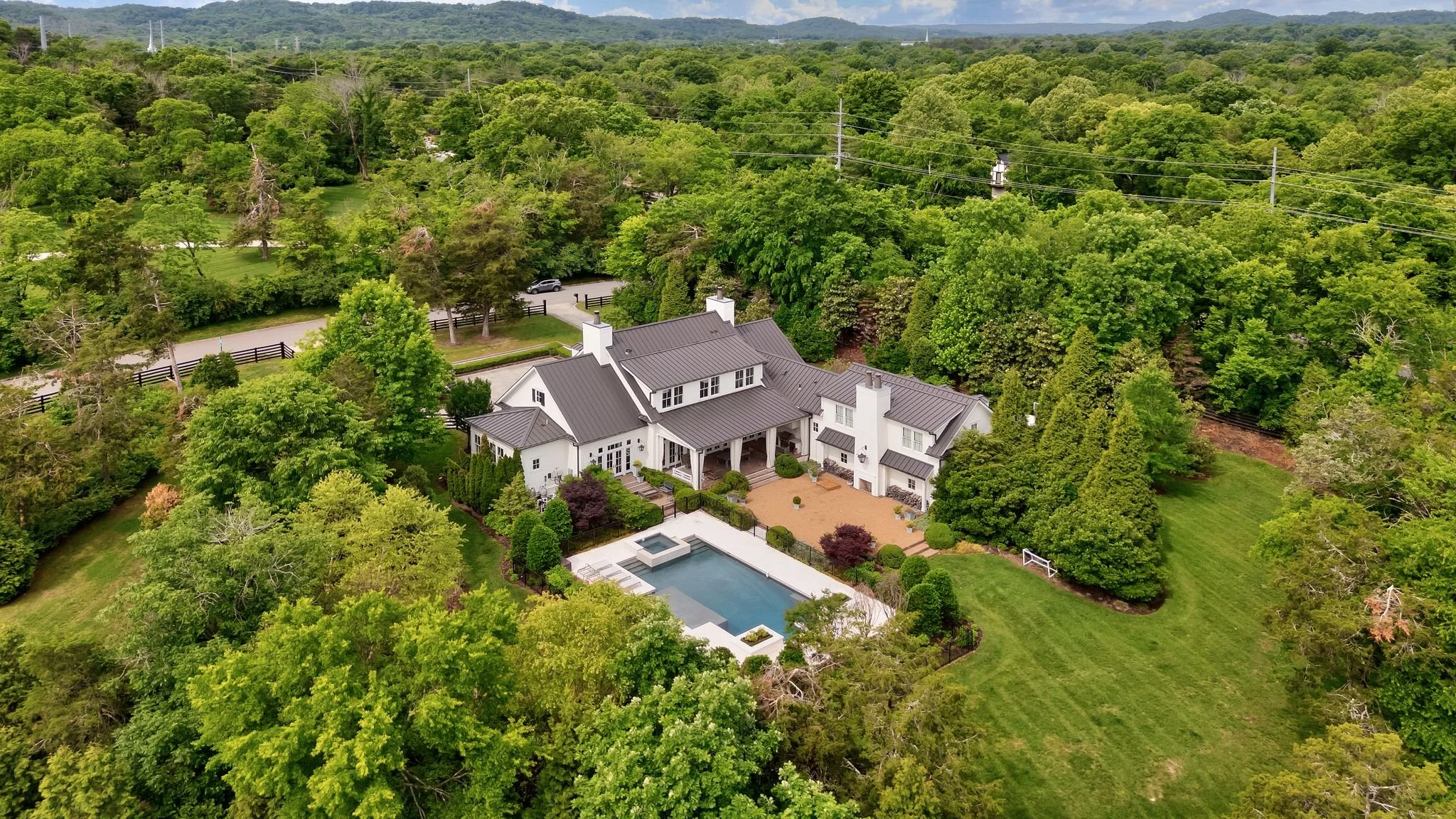 A large white house surrounded by lush green trees with a backyard pool and patio area, set in a scenic, wooded landscape with hills in the background.