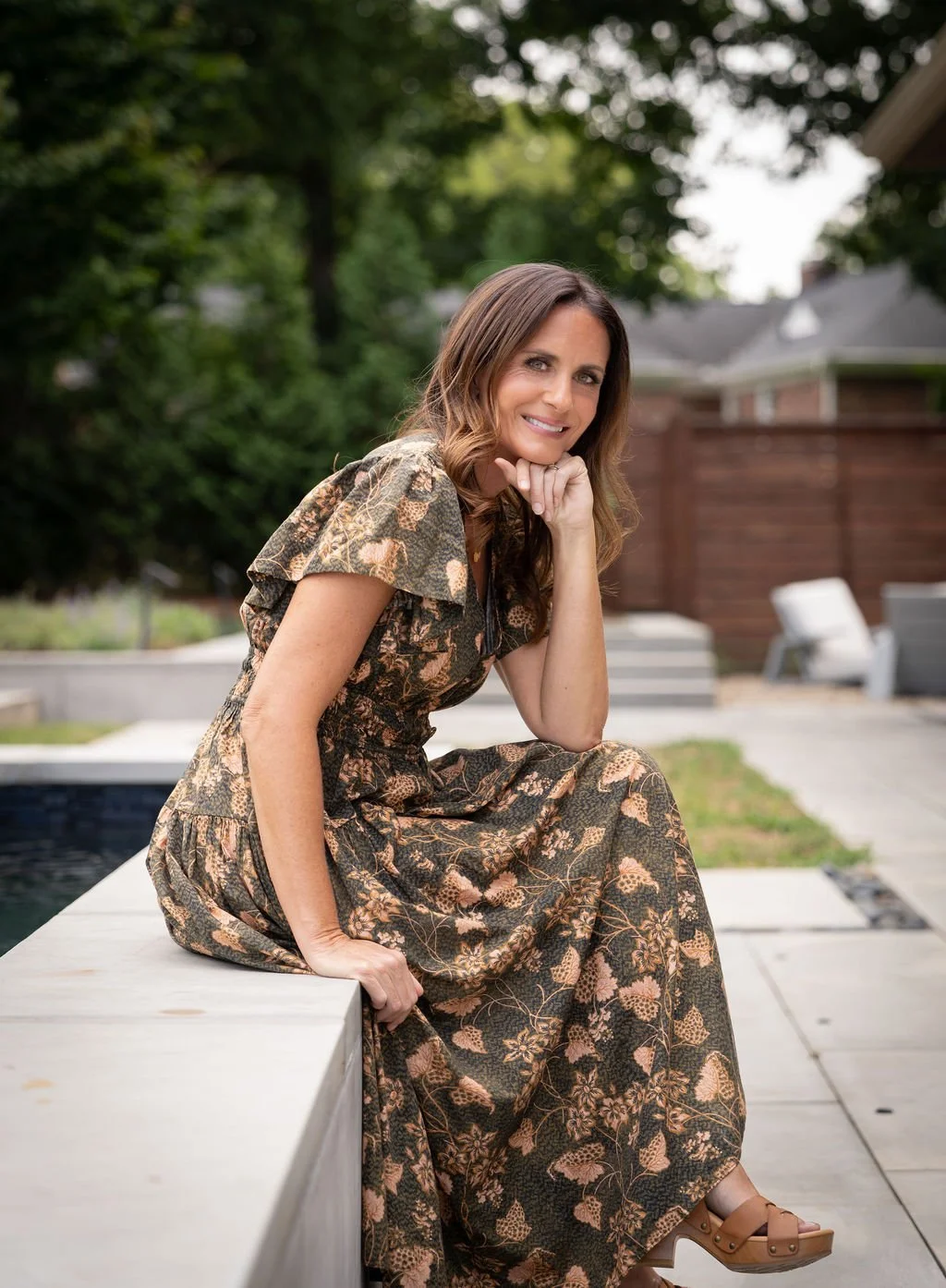 Brittany sits on concrete ledge outdoors, wearing a patterned dress, smiling, with greenery and a backyard in background.