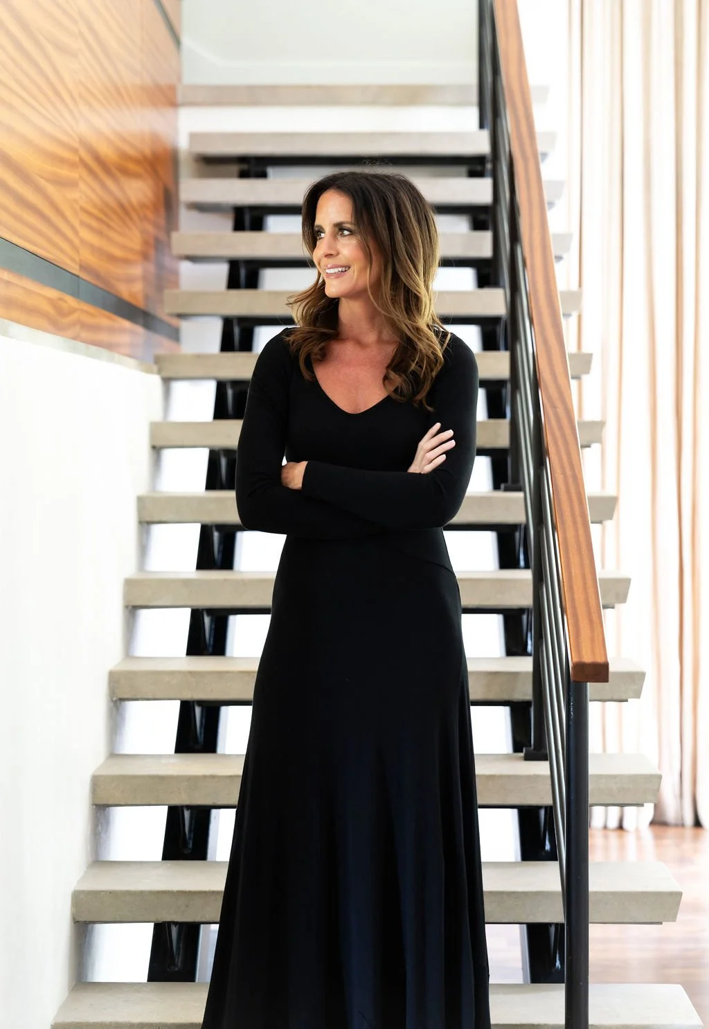 Brittany Sircy stands on a staircase with crossed arms, smiling, wearing a long black dress, in a modern interior with wooden and glass elements.