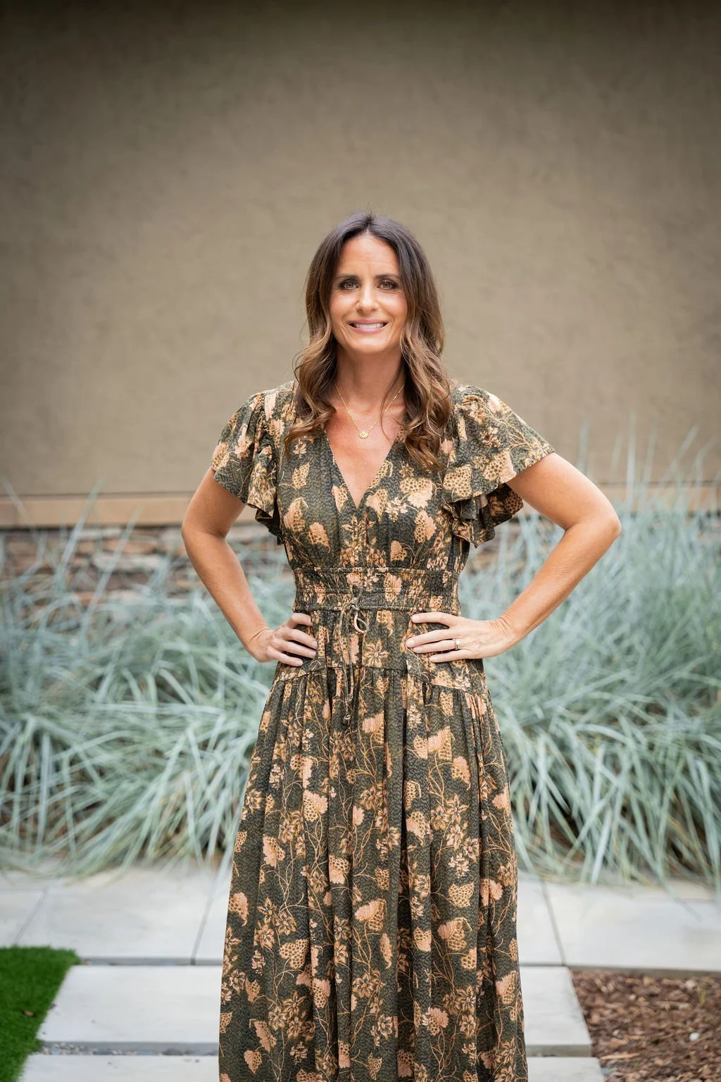 Brittany wears a floral print dress standing outdoors with hands on her hips, smiling, in front of greenery and a beige wall.