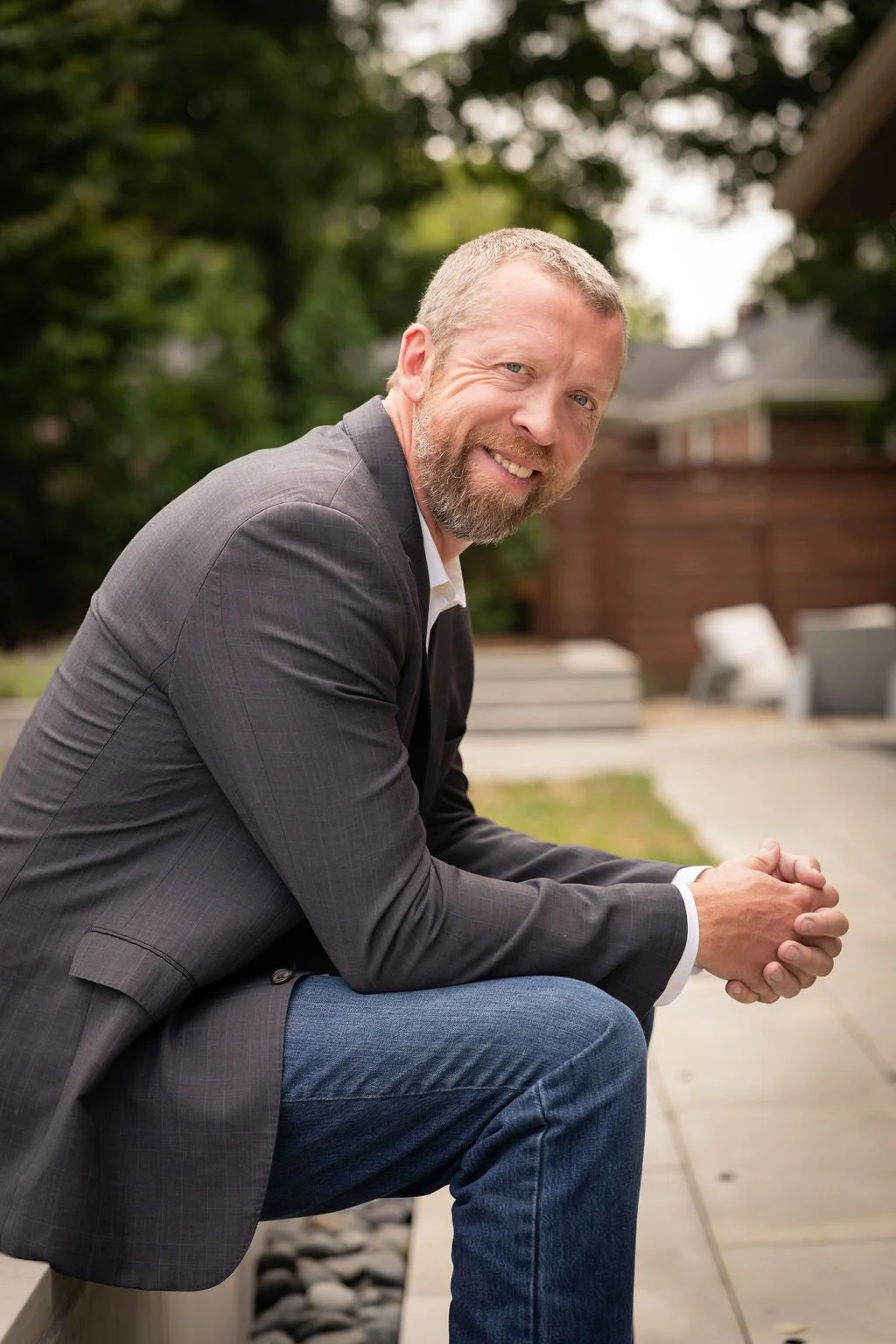 Josh wears a black suit jacket and jeans, sitting outdoors on a bench and smiling at the camera.