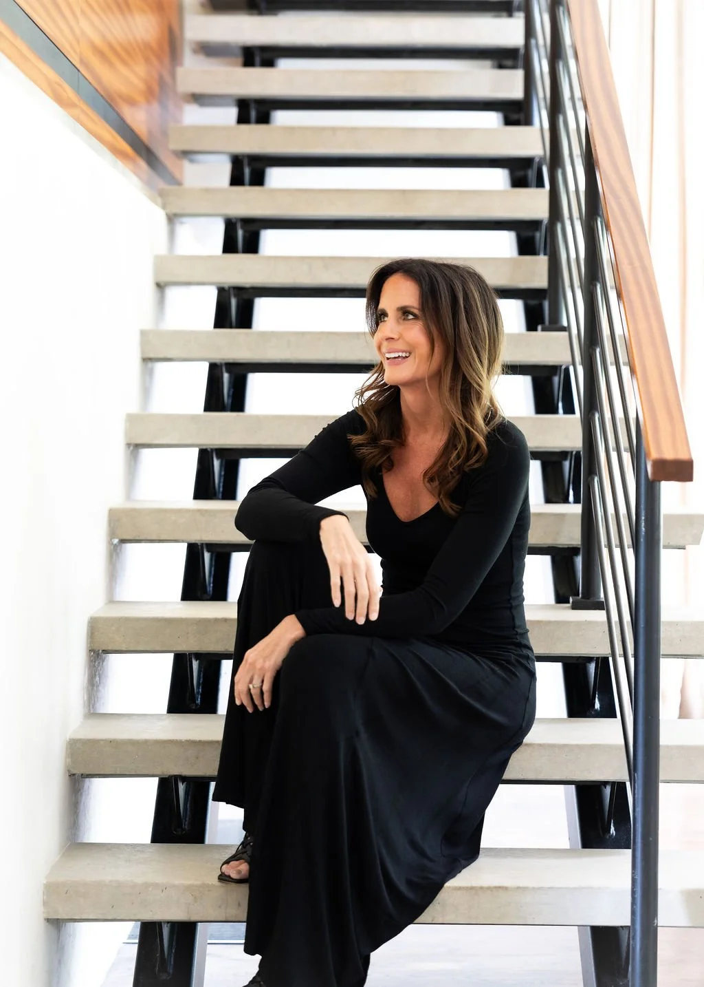 Brittany wears a black long-sleeve dress sitting on a modern staircase, smiling and looking to her right, with a wooden handrail and metal railing.
