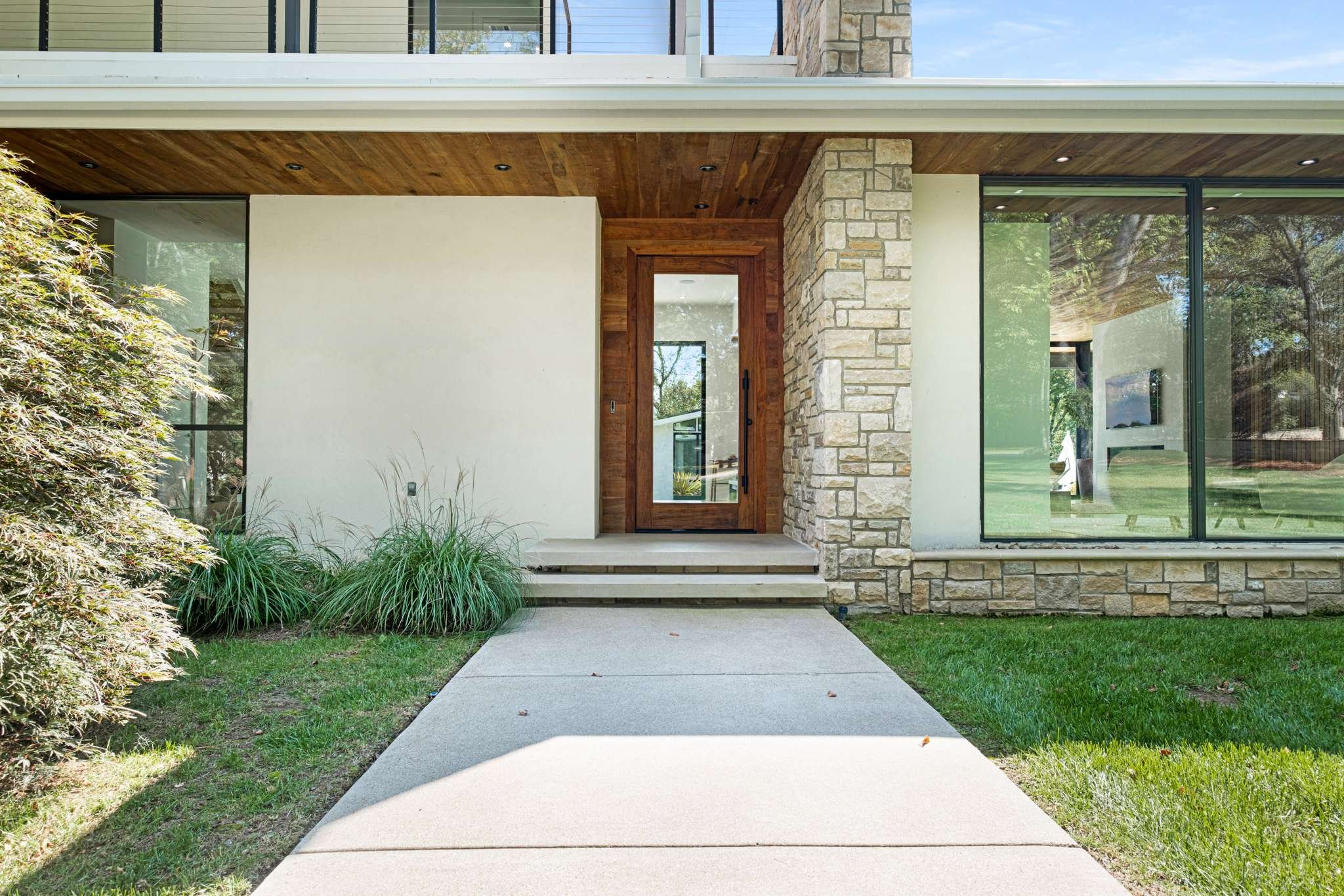 Front entrance of a modern house in Tennessee with a concrete pathway, green lawn, bushes, stone and wood exterior, large glass windows, and a wooden door.