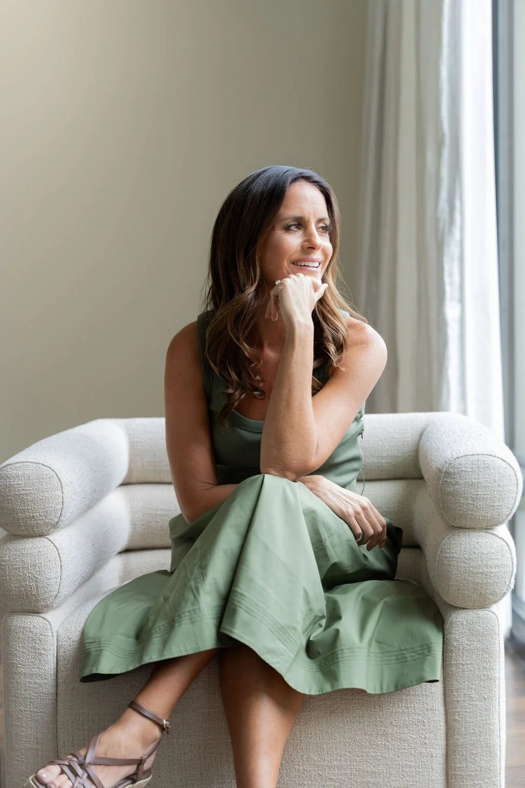 Brittany sits on a white couch by a window, smiling and looking to her right, wearing a green dress and sandals.