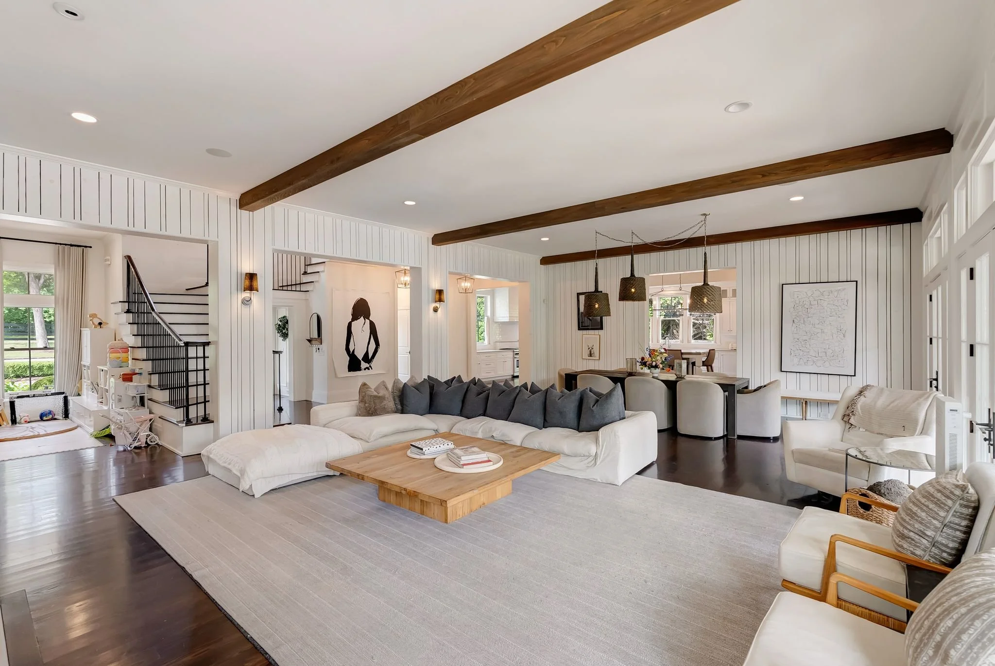 Bright living room with white panel walls, wooden beams on the ceiling, a large white sofa with dark throw pillows, a wooden coffee table with books, and artwork on the walls. Multiple windows and pendant lights illuminate the space.