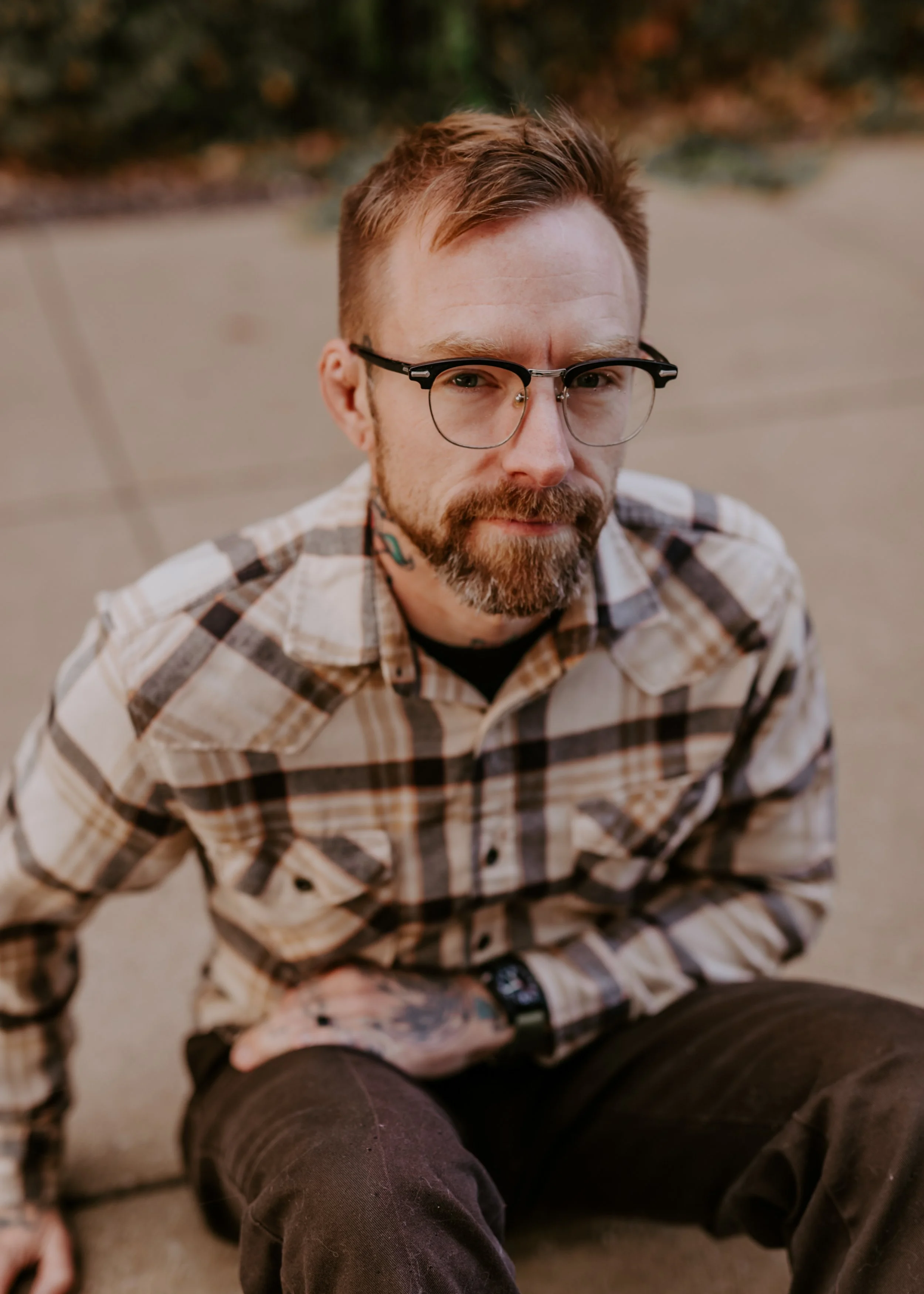 Close-up of a man with short reddish hair and glasses, wearing a plaid flannel shirt, sitting outdoors on the ground with other people, with blurred greenery in the background.