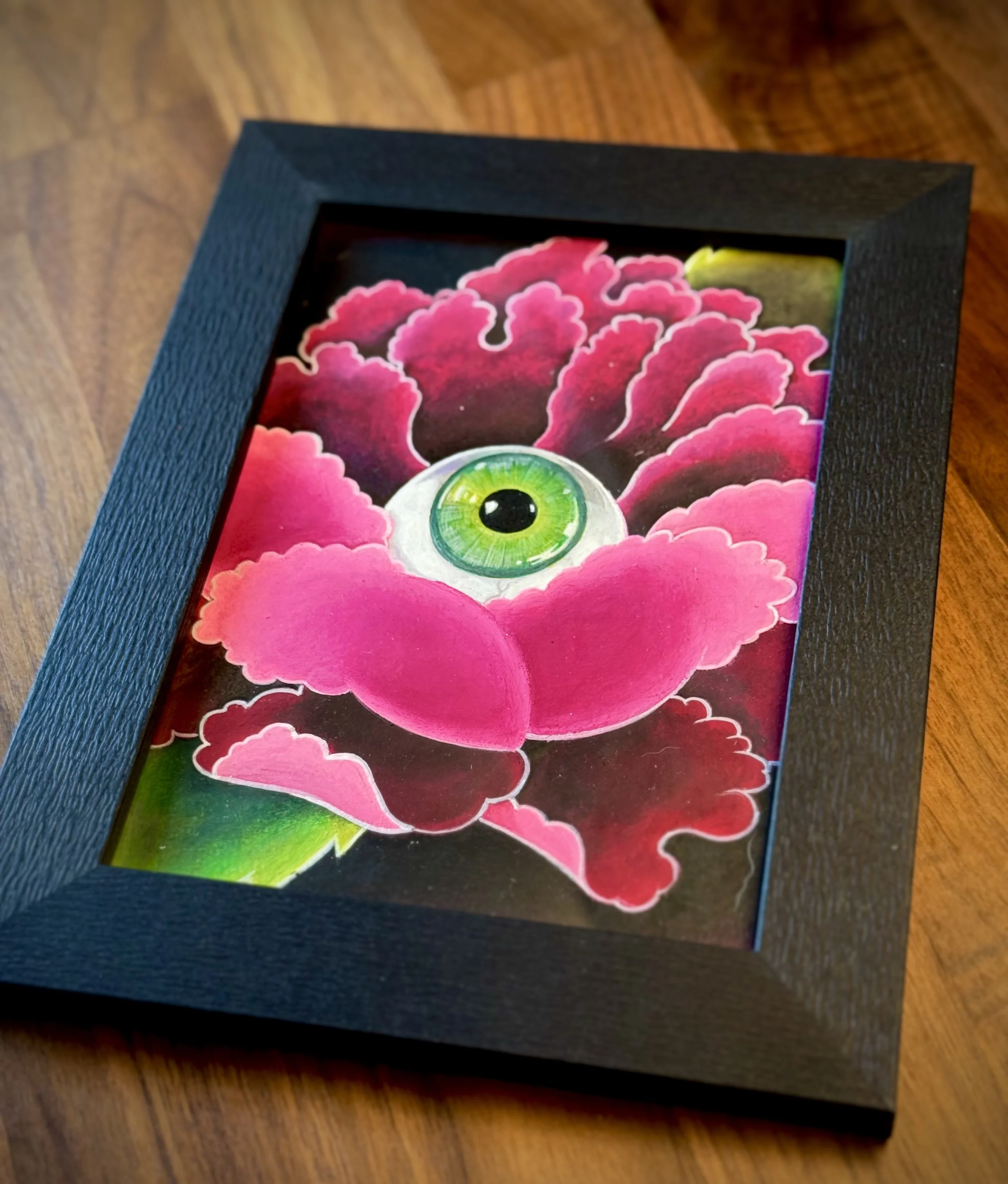 Framed artwork of a peony flower with a green eye in its center, placed on a wooden surface.