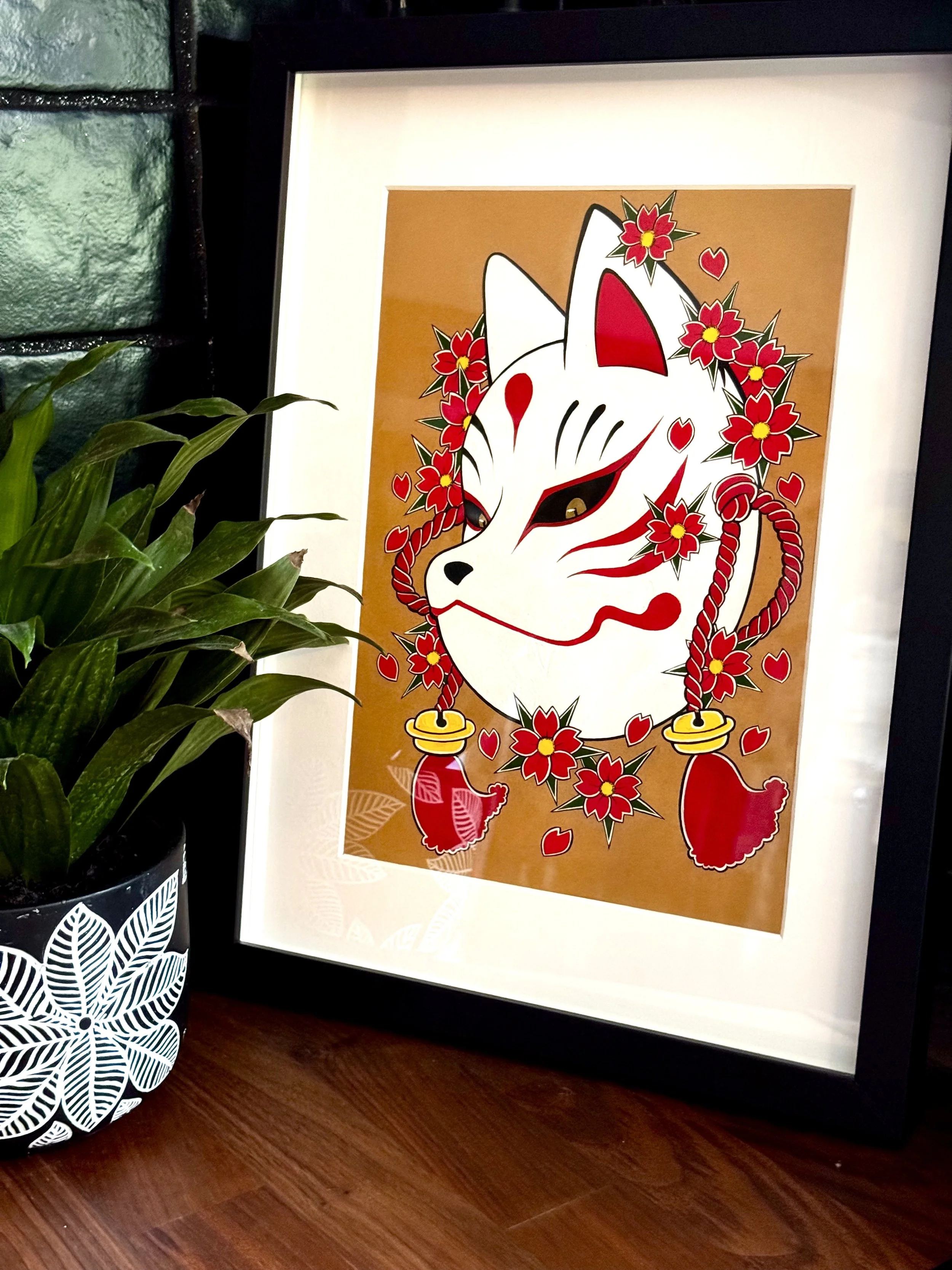 Framed artwork of a white fox mask with red and black markings, surrounded by cherry blossoms and gold bells, on a brown background.