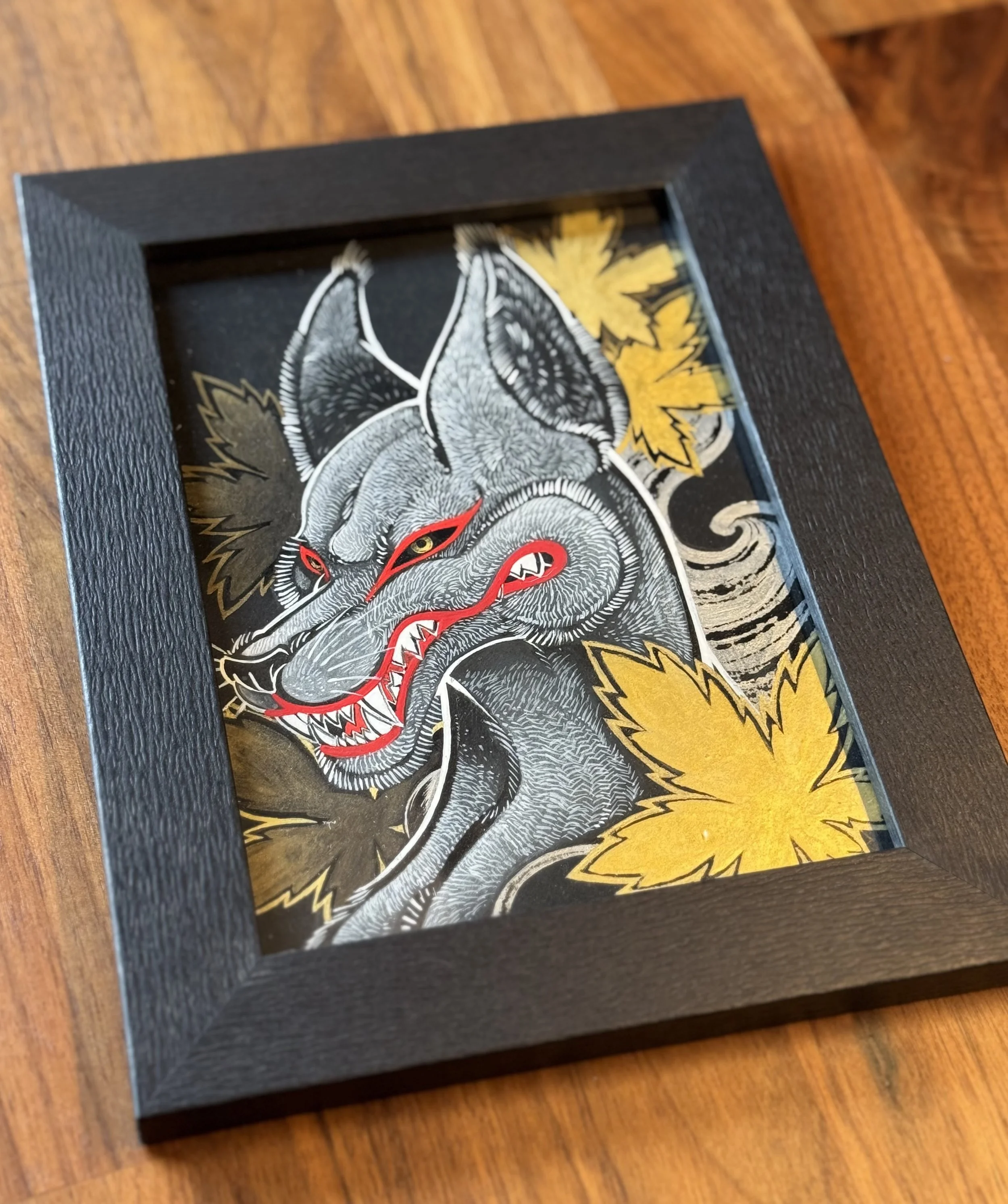 Close-up of a framed artwork featuring a detailed, nogitsune with red eyes, set against a background with golden maple leaves, placed on a wooden surface.