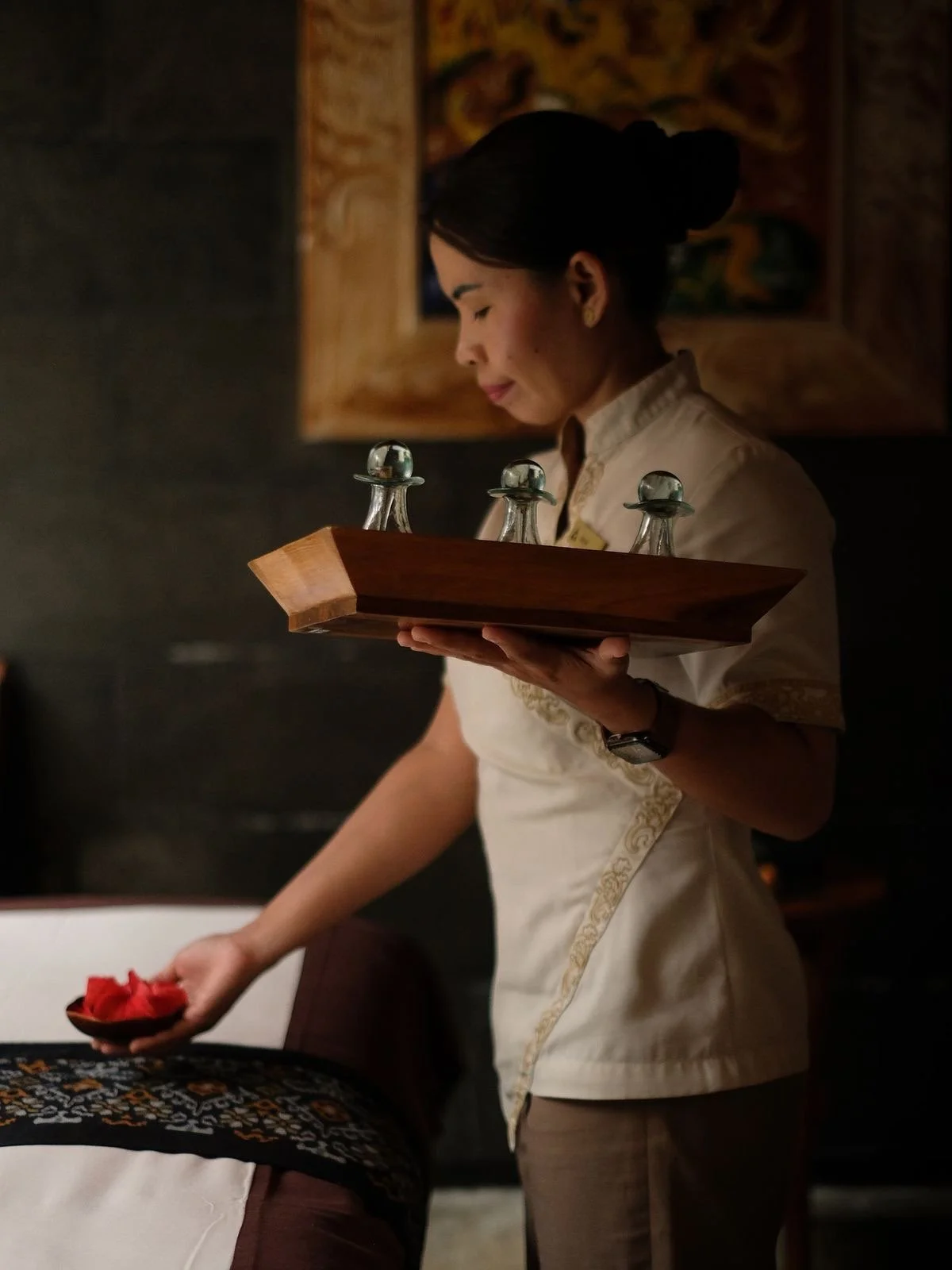 A woman dressed in traditional attire is holding a tray with glass teardrop-shaped bottles on it, while serving food on a plate, in a dimly lit room with artwork on the wall in the background.