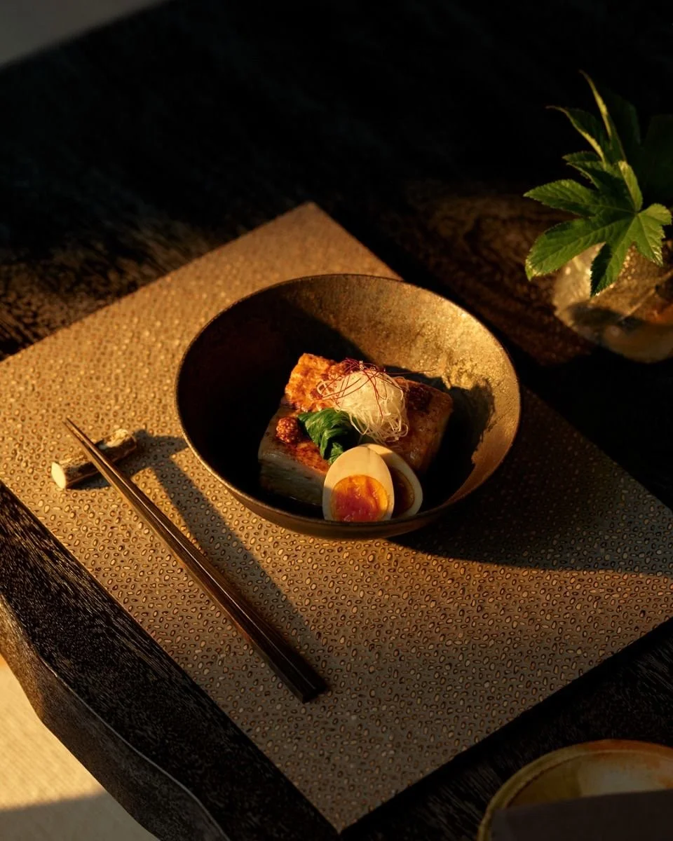 A black bowl of Japanese-style food with sliced boiled egg, topped with green leaves and thin red strands, placed on a textured beige placemat, with chopsticks beside it, and a plant in the background.