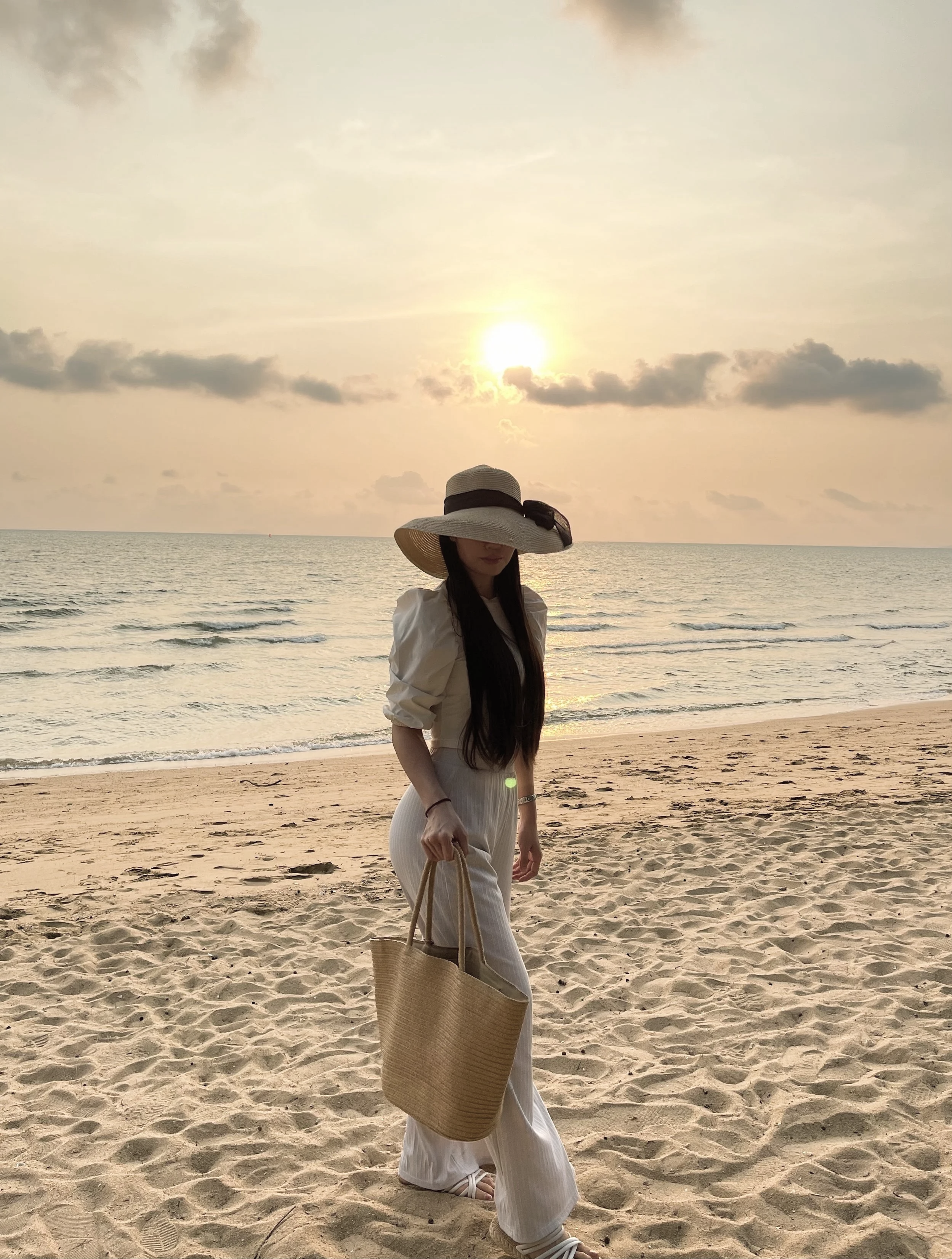 A woman in a white dress and wide-brimmed hat walking on the sandy beach at sunset, carrying a large straw tote bag.
