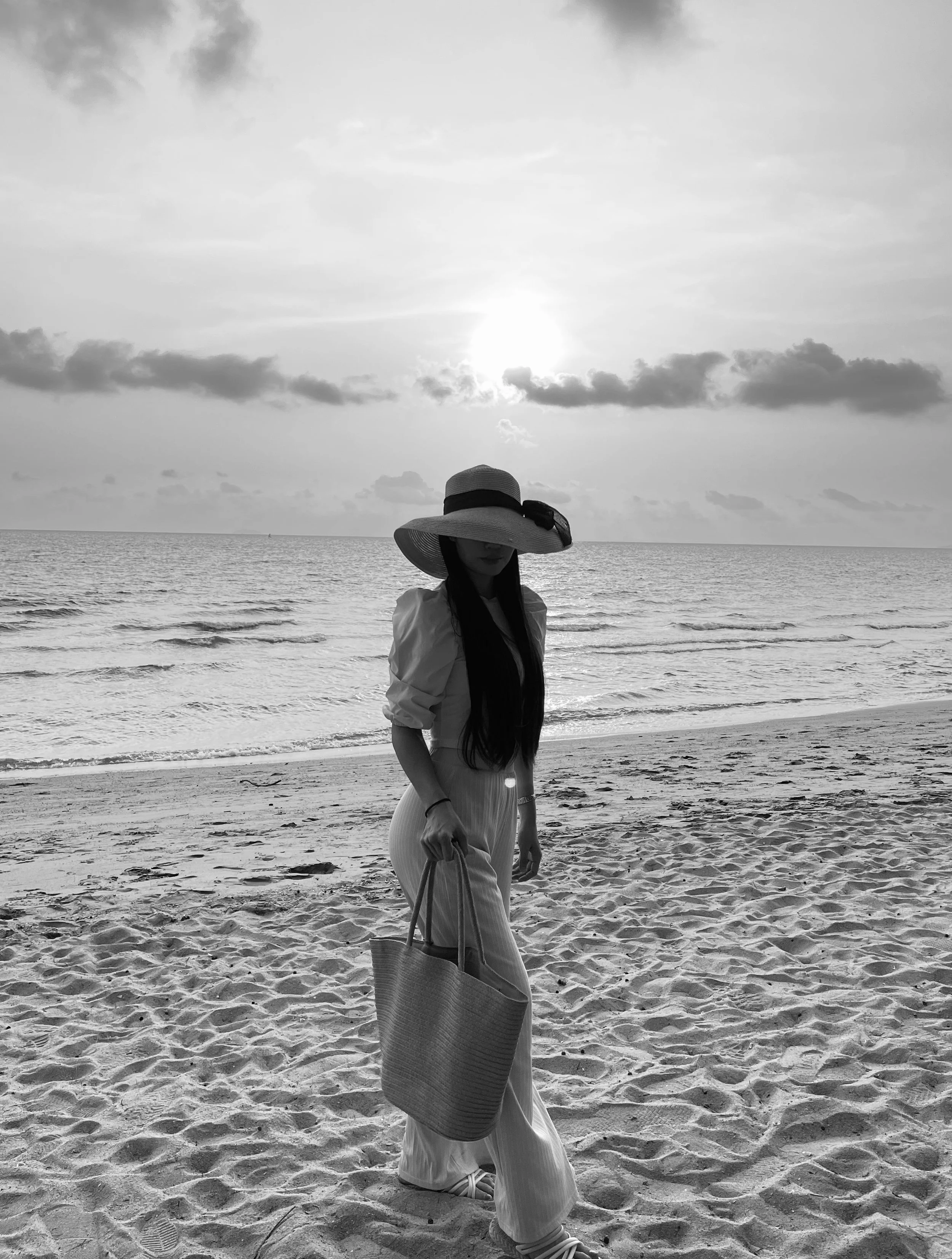 A woman standing on a sandy beach with ocean waves behind her, wearing a wide-brimmed hat, a long-sleeved top, pants, and holding a large woven bag, with the sun setting or rising in the sky and clouds above.