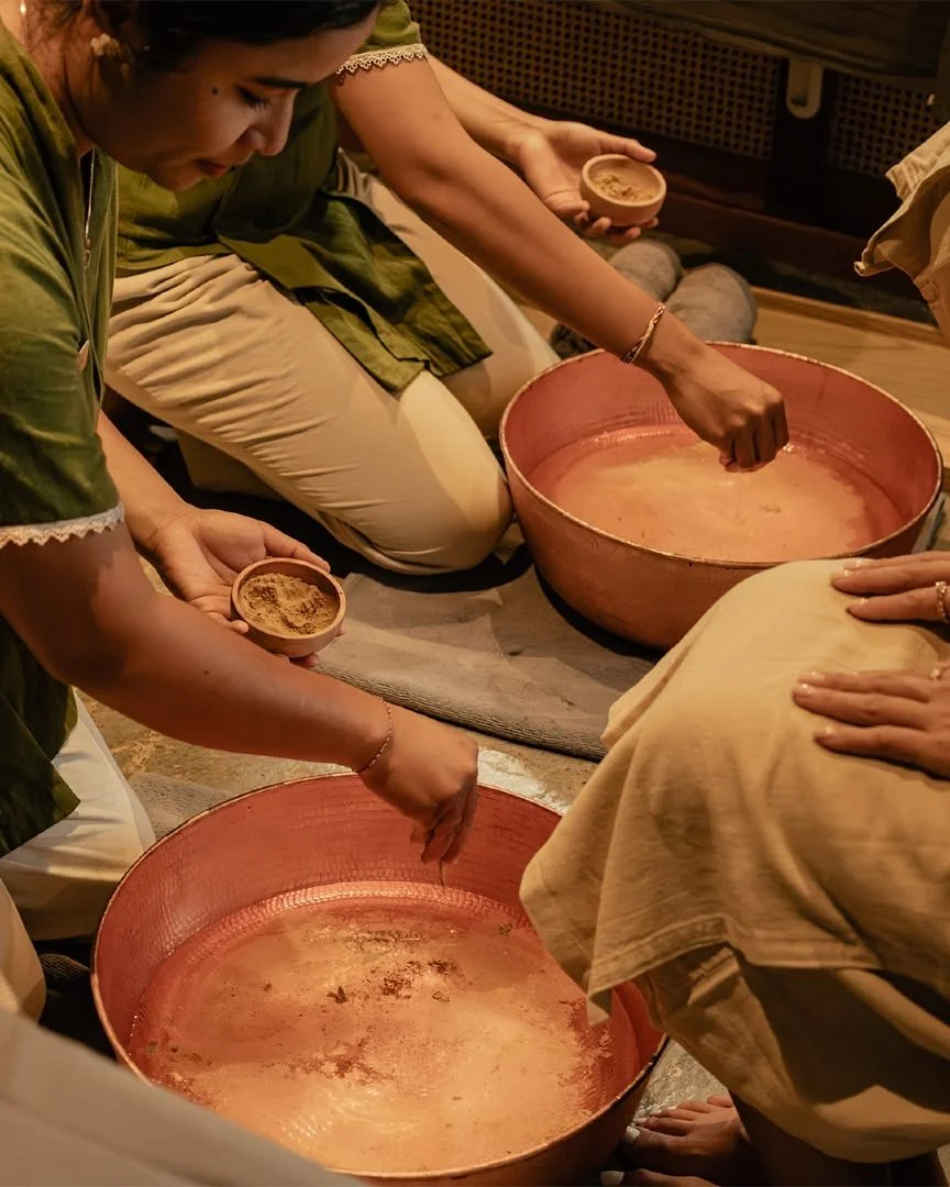 People sitting in a circle, performing a traditional cleansing ritual with bowls of water and powders on the floor.