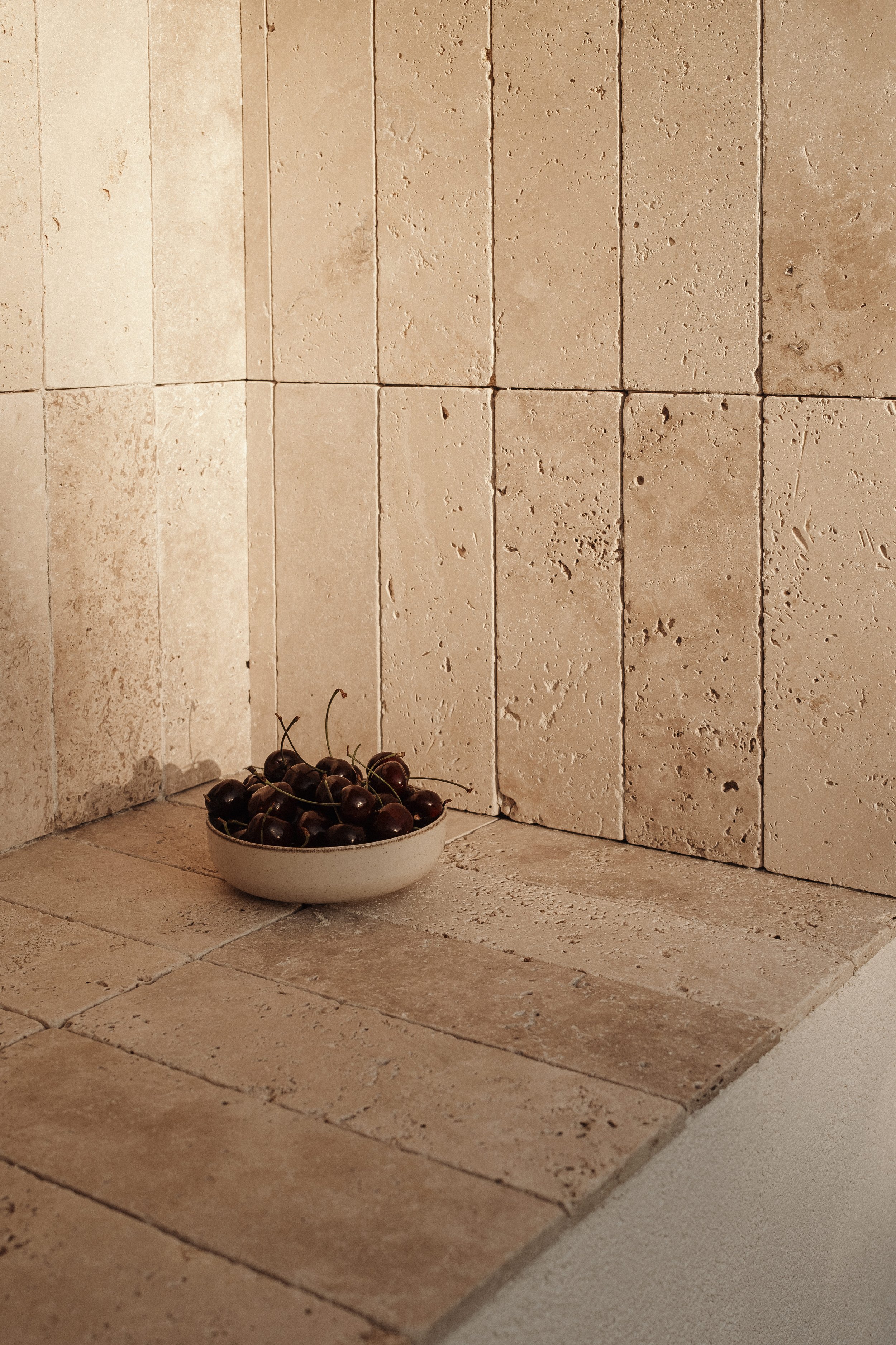 A beige ceramic bowl filled with cherries placed on a tiled piedra stone surface in front of a stone wall.