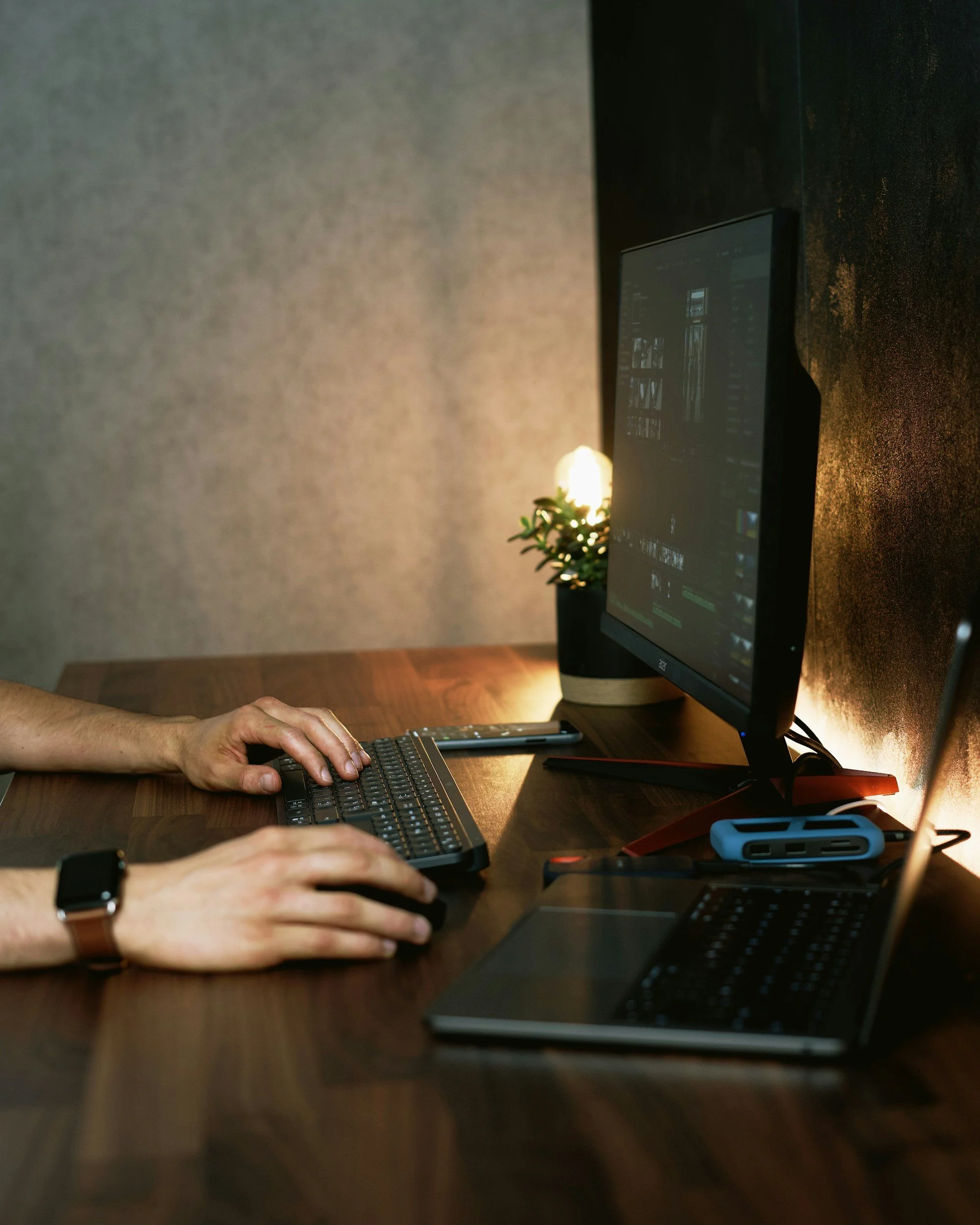Deux personnes travaillent sur un ordinateur à un bureau en bois, avec un deuxième clavier et d'autres accessoires technologiques. La pièce est tamisée avec une lumière douce et une plante en arrière-plan.